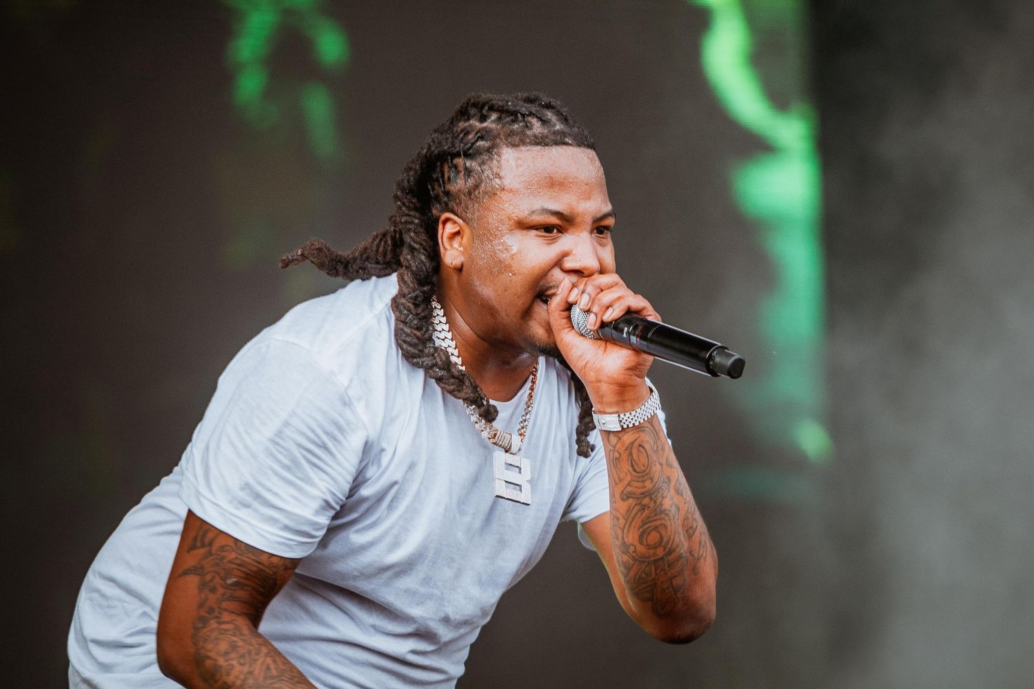 Male rapper performing on stage in a white t-shirt with dreadlocks and a chain necklace outdoors.