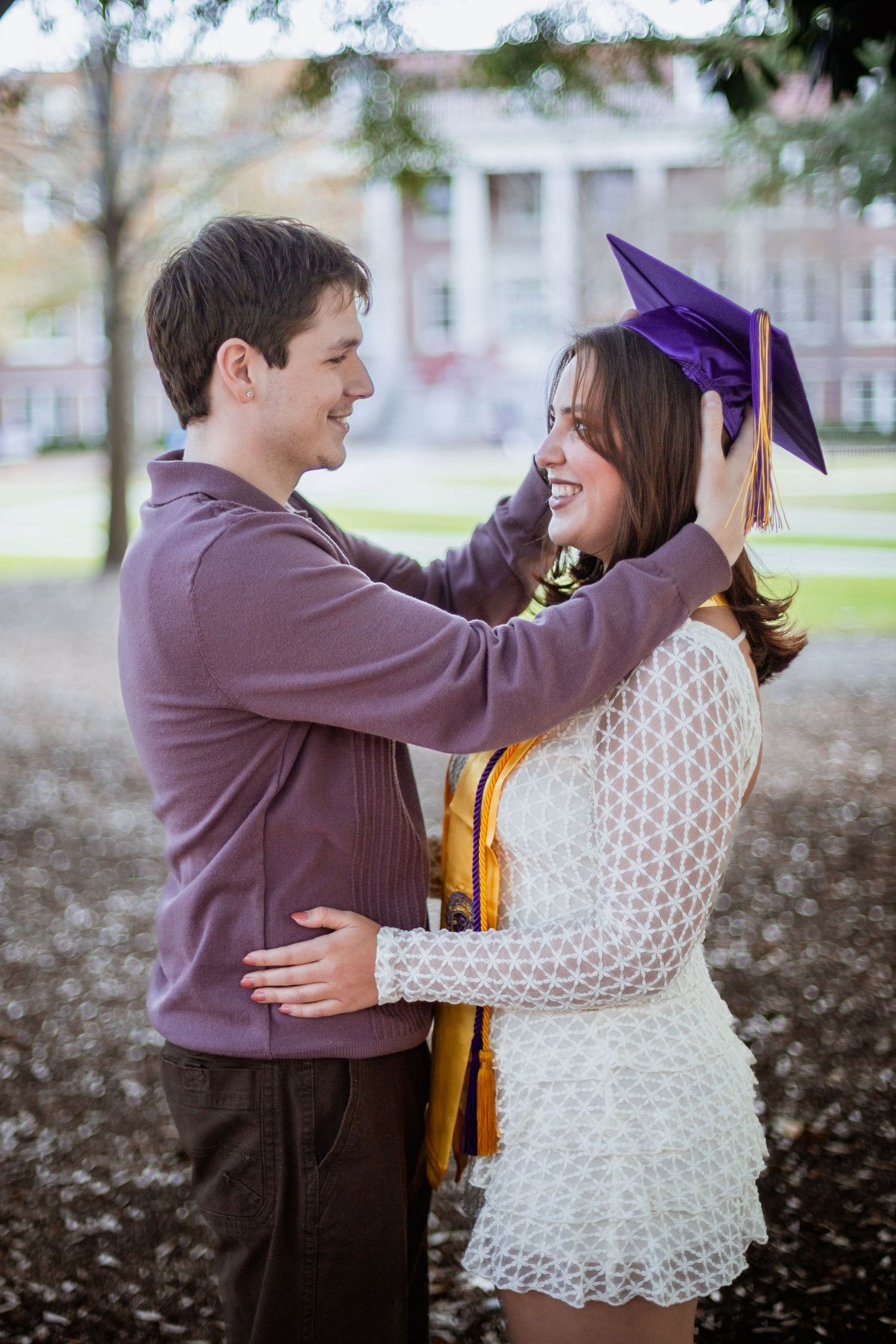 Young couple smiling outdoors as man places purple graduation cap on womans head near university building.