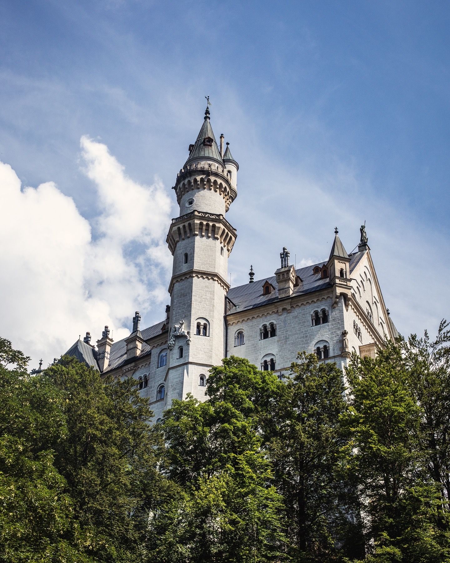 Neuschwanstein Castle tower rises above lush green trees against a blue sky with white clouds.