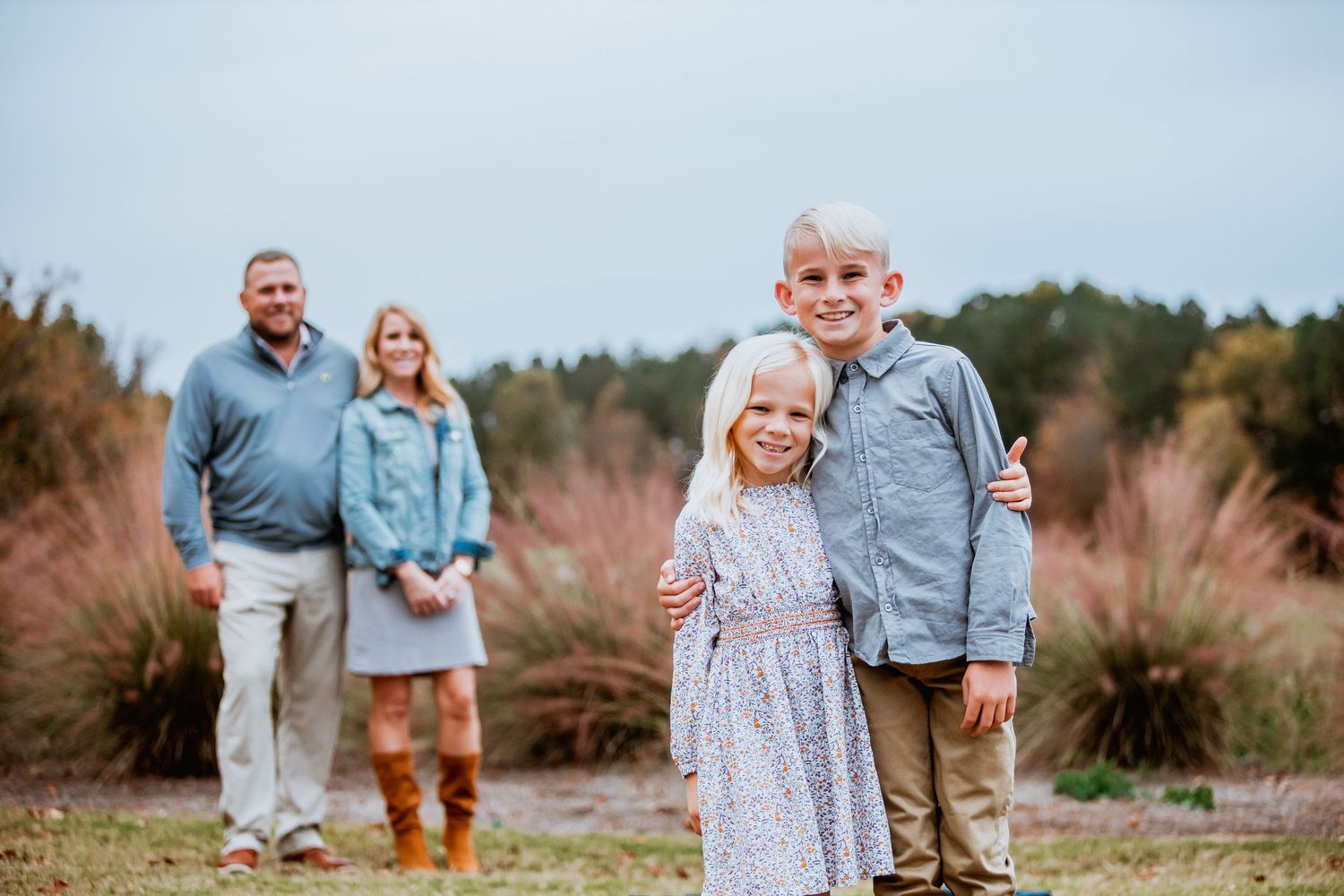 Family portrait with two smiling children in foreground and parents standing in colorful autumn field.