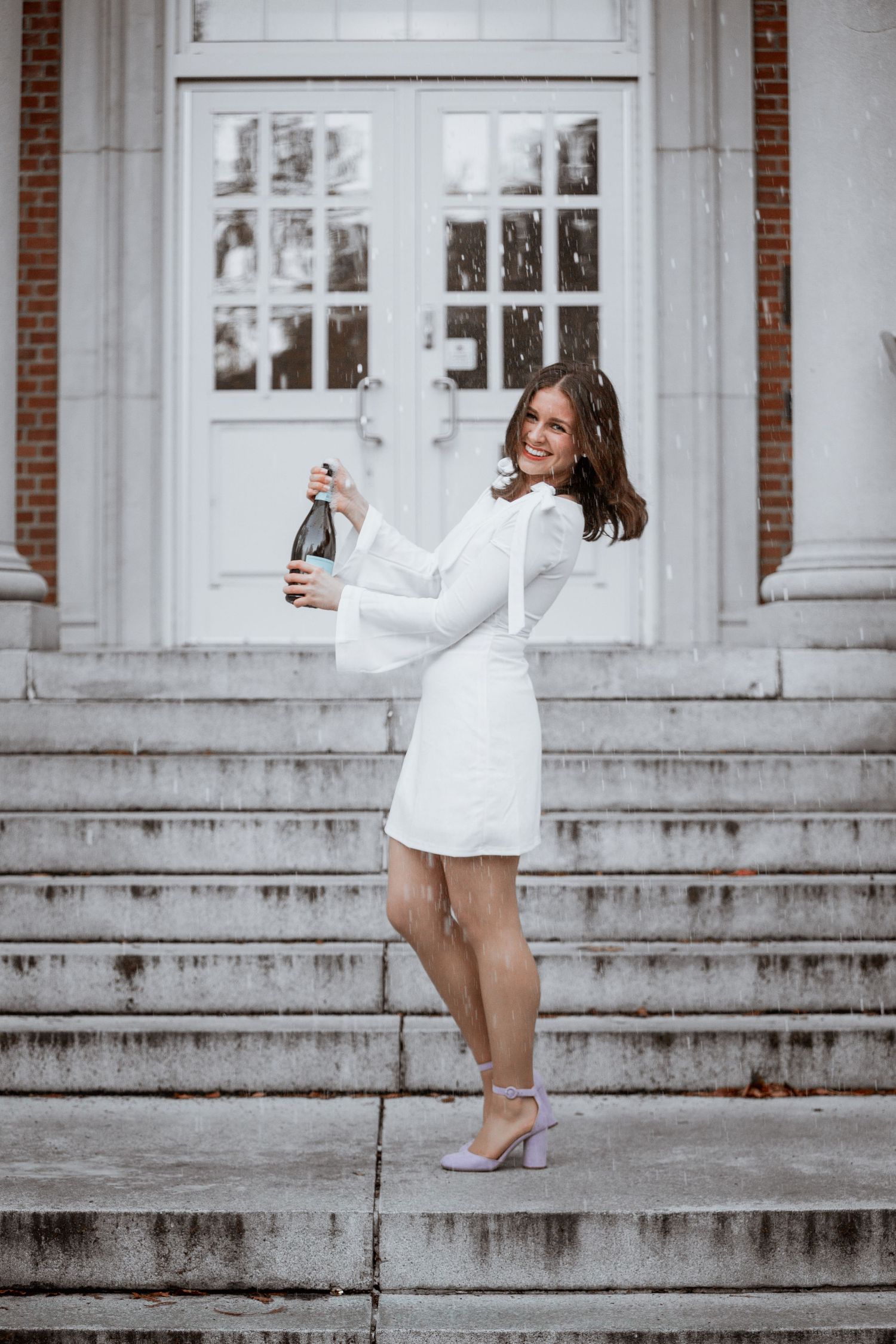 Young woman in white mini dress popping champagne bottle on steps of elegant building with white doors.