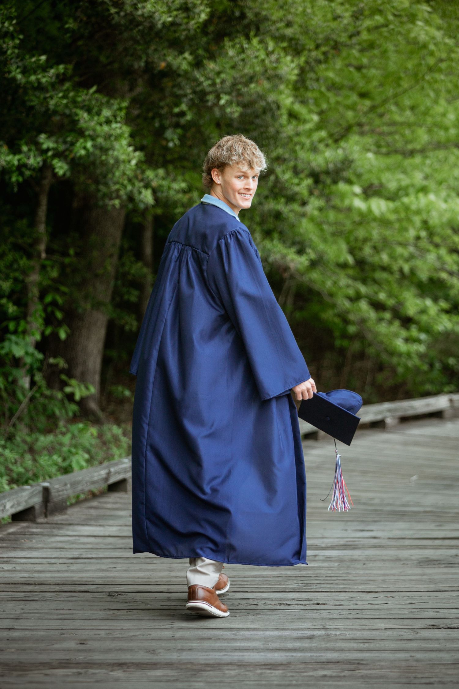 Graduate in blue cap and gown walks along wooden boardwalk path surrounded by lush green trees.