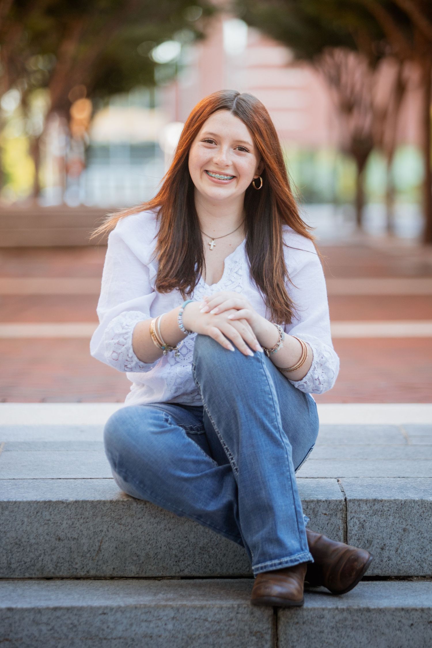 Smiling teenage girl with red hair sitting on stone steps outdoors in white top and jeans.
