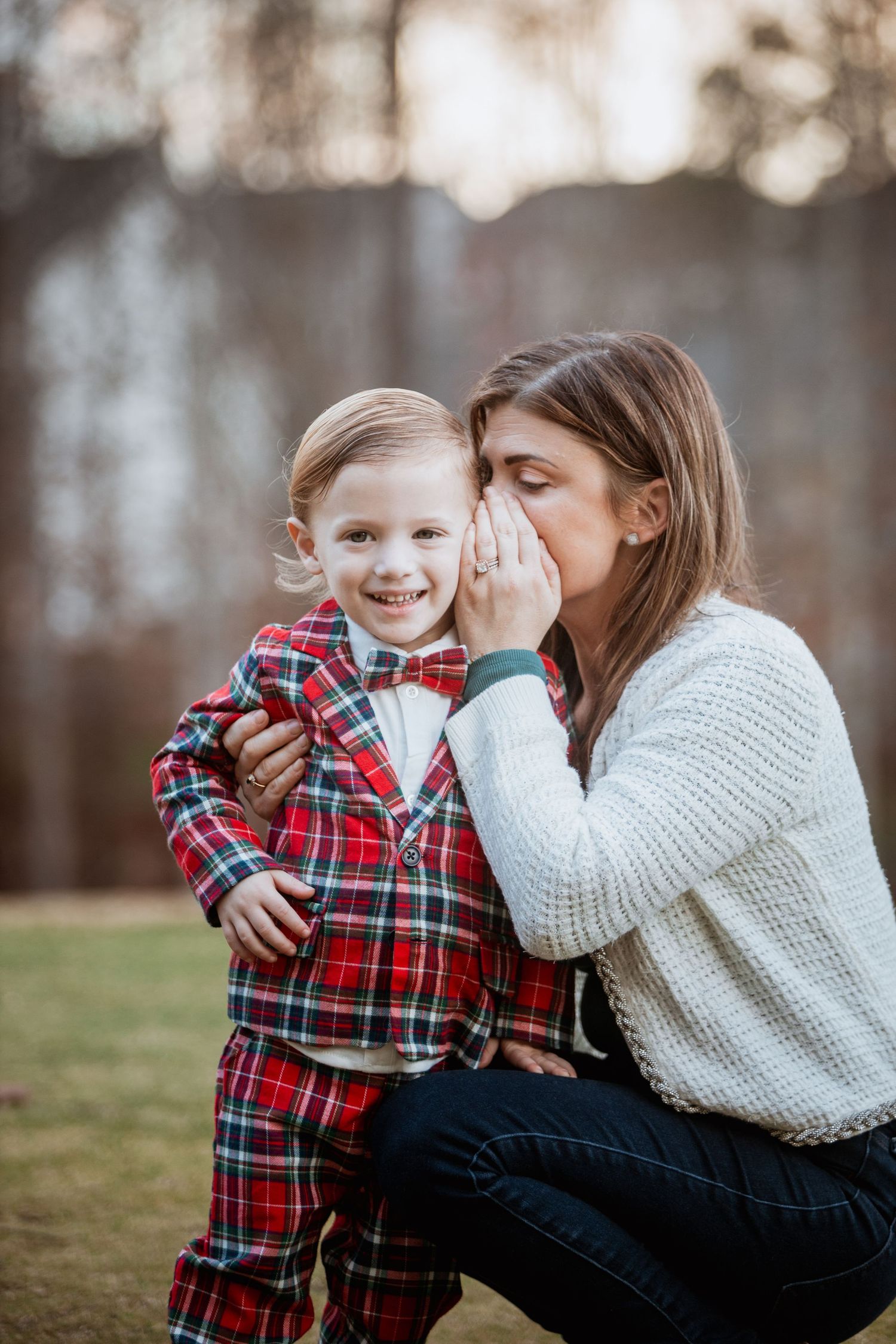 Woman whispering to smiling toddler boy in red plaid suit outdoors.