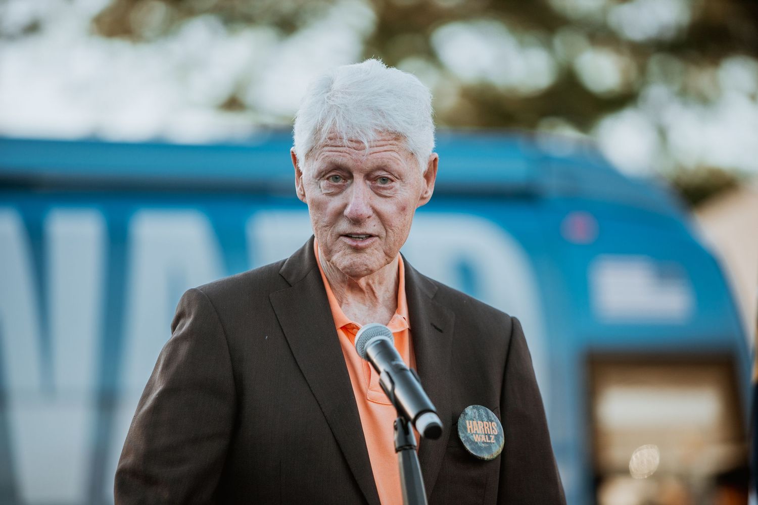 Elderly man with white hair speaking at a microphone outdoors, wearing a dark blazer and orange shirt with a campaign button.