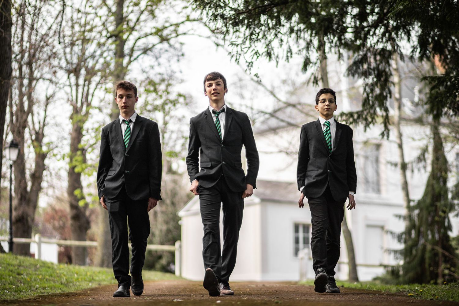 Three teenage boys in dark suits with green striped ties walk confidently along a tree-lined path.