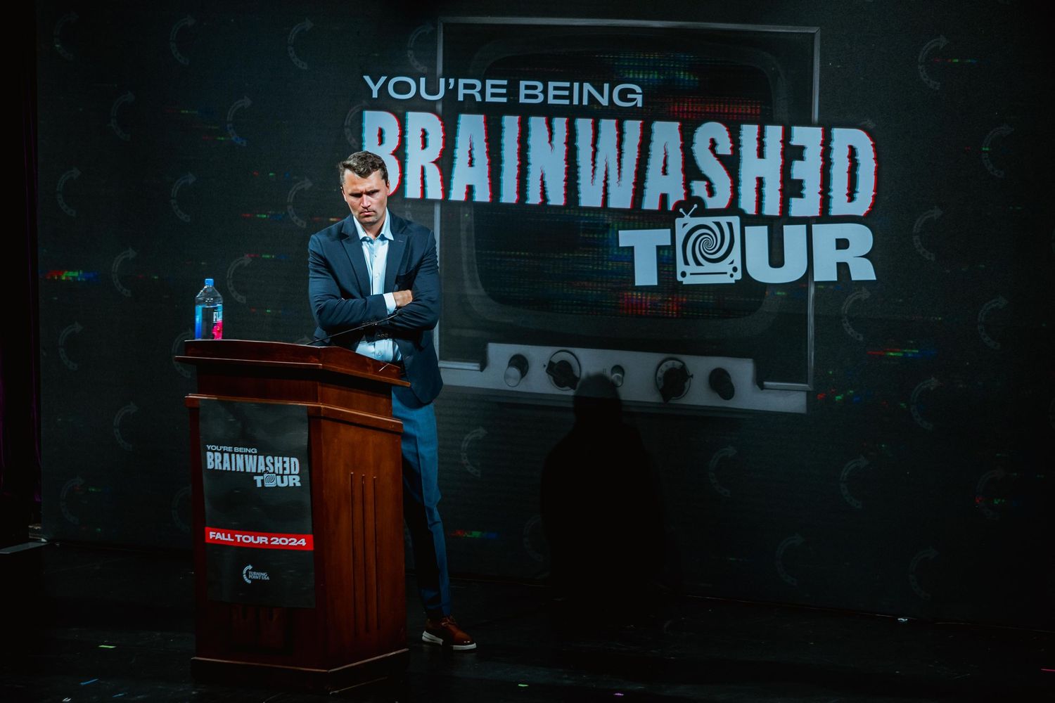 Man in suit presenting at You're Being Brainwashed Tour event on stage.