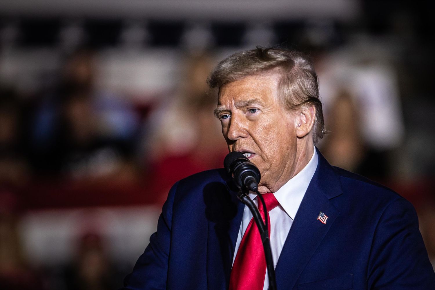 Donald Trump speaks passionately into a microphone at a political campaign rally wearing a blue suit and red tie.