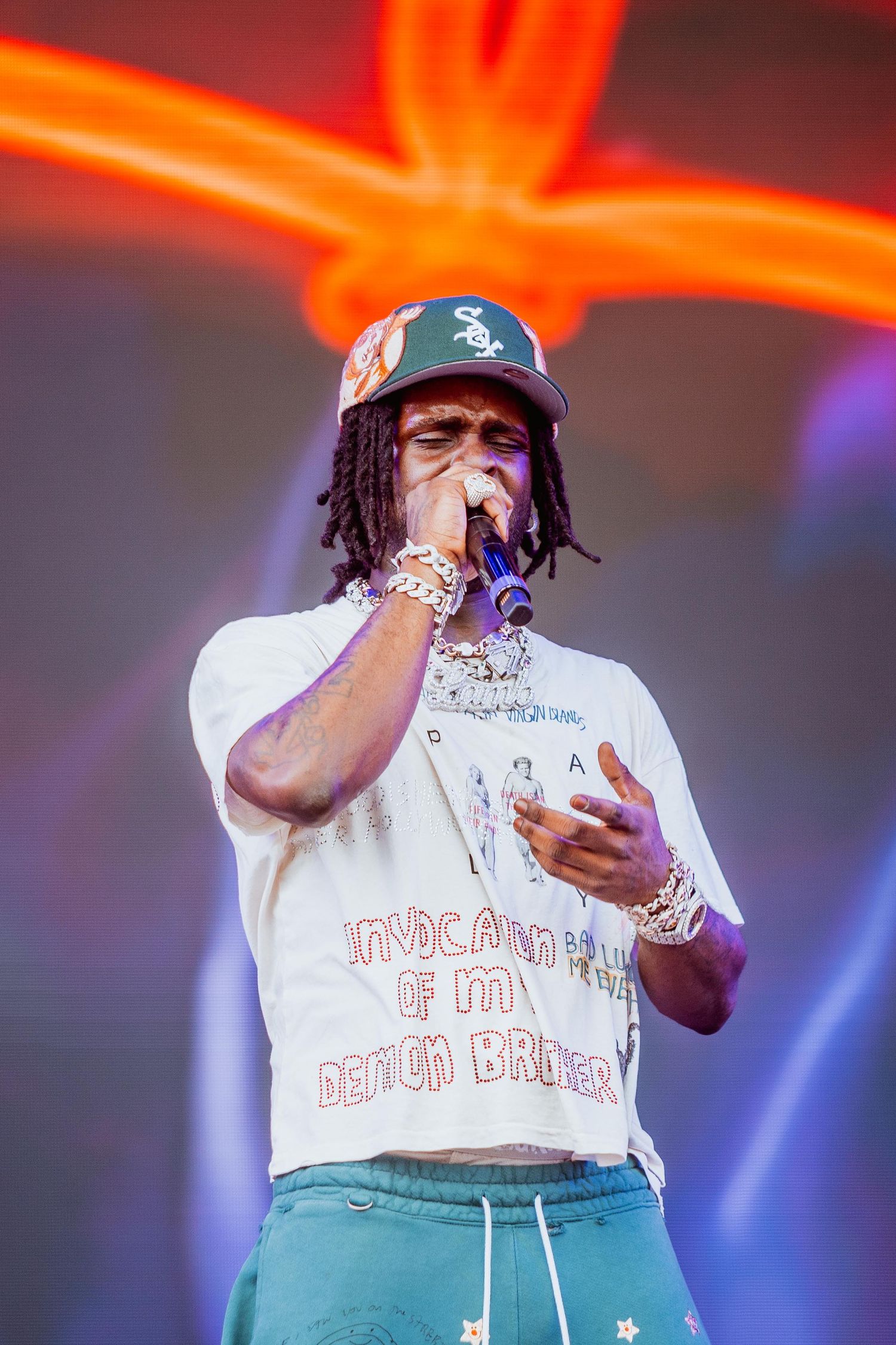 Rapper performing on stage wearing a white graphic tee, teal pants, chains, and a baseball cap under neon lights.
