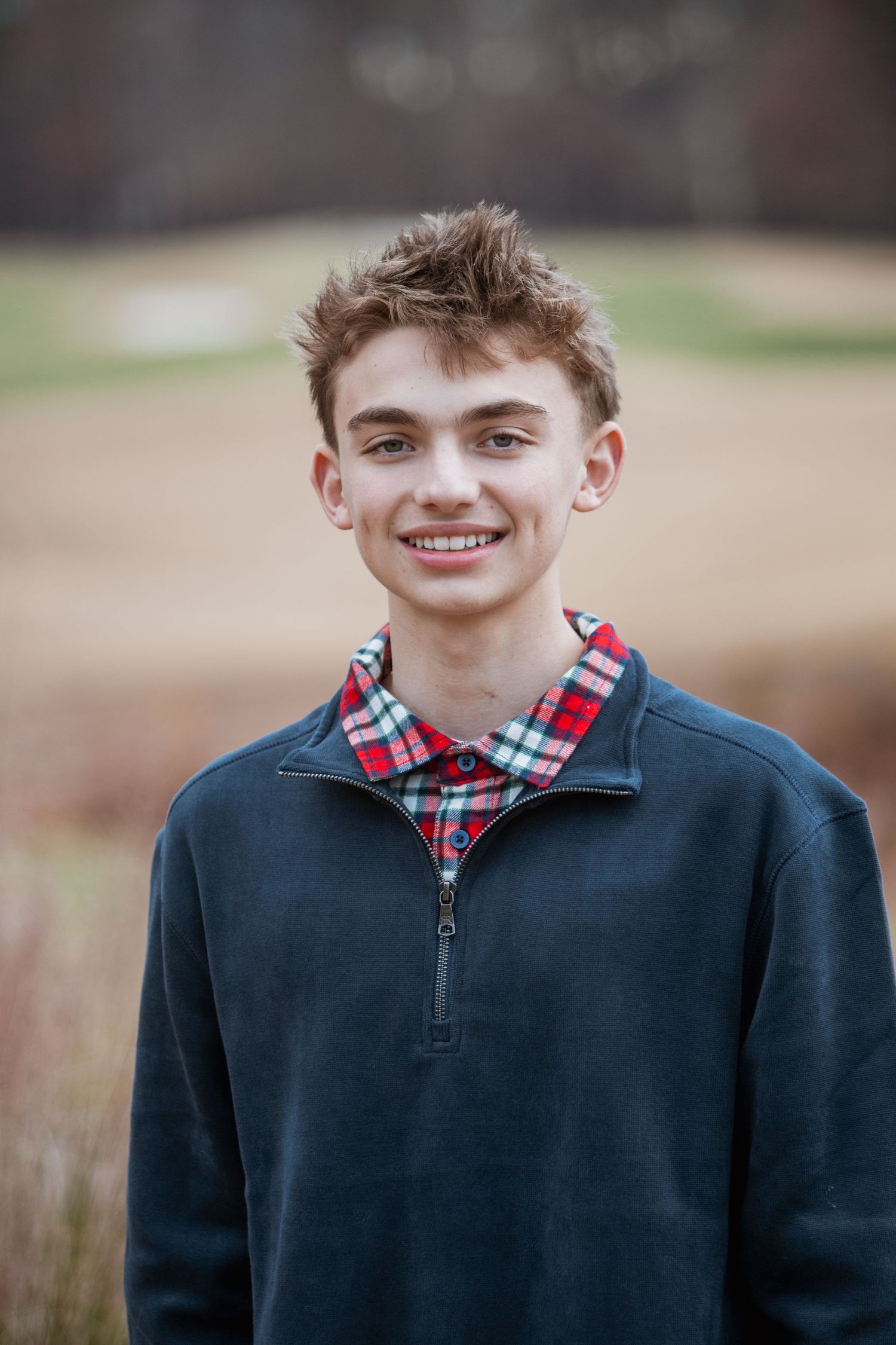 Smiling teenage boy in navy pullover with plaid collar shirt posing outdoors.