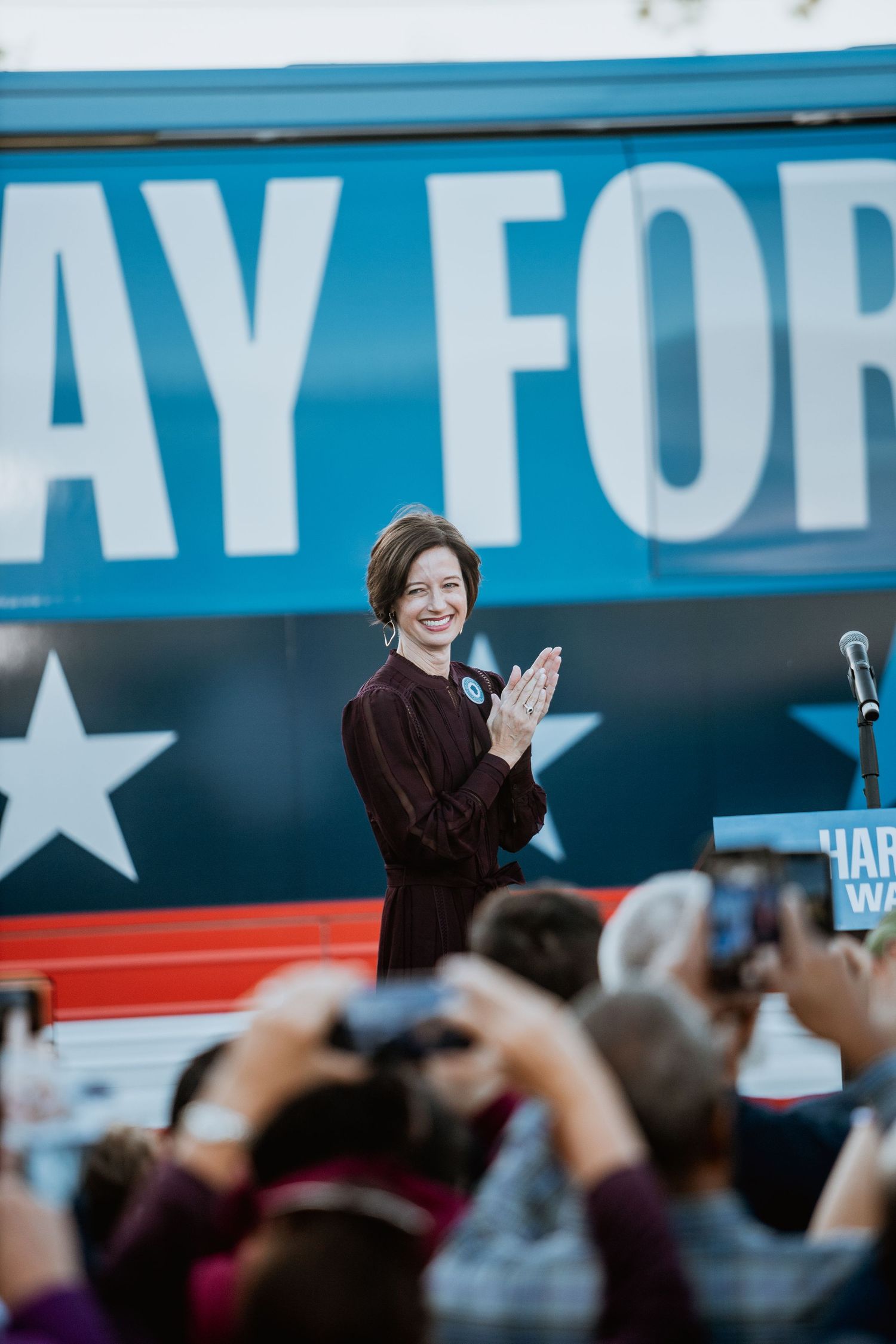 Woman in burgundy dress clapping at political campaign rally in front of blue campaign bus.
