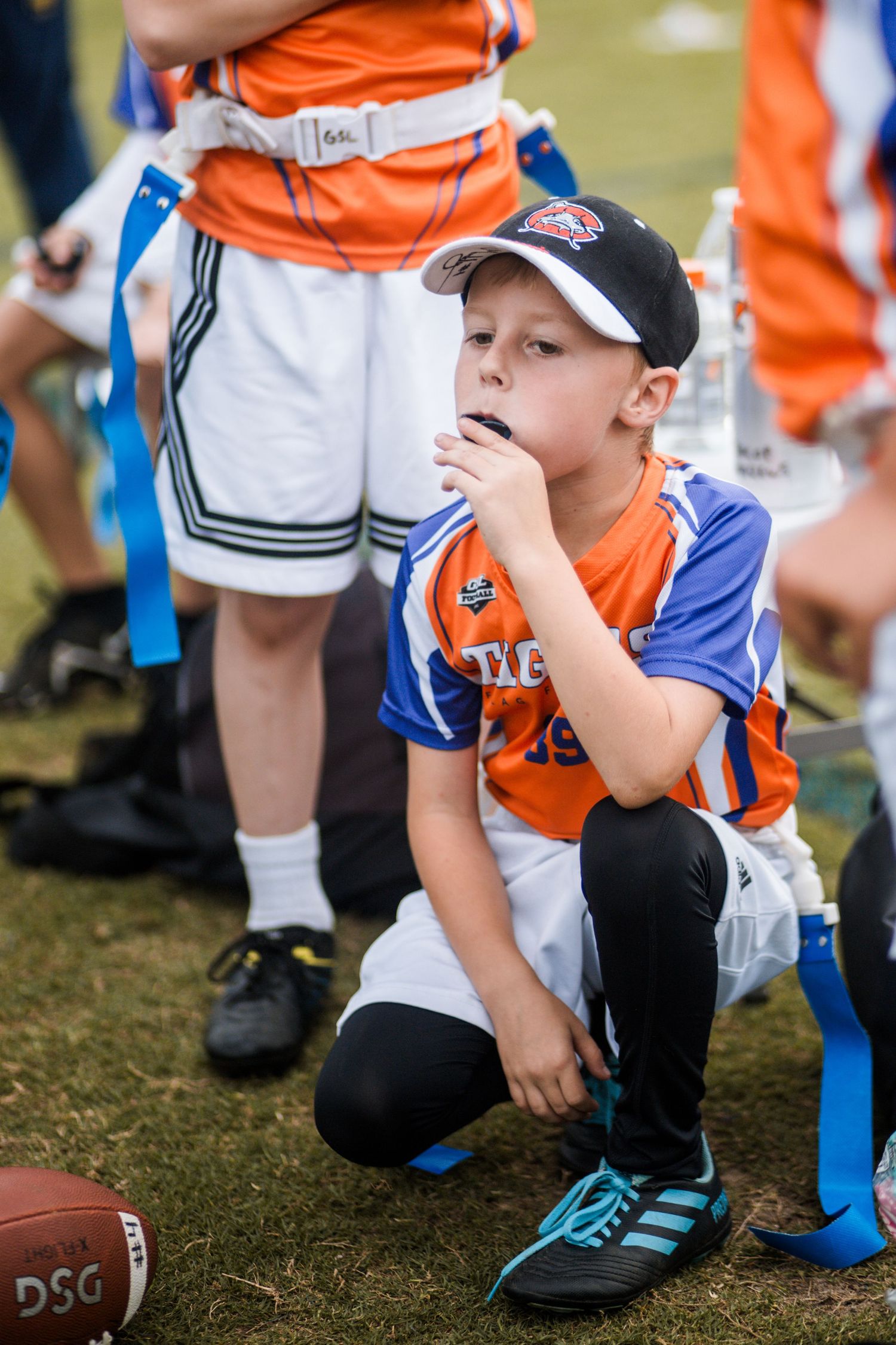 Young boy in Tigers flag football jersey kneeling and drinking water on sideline.