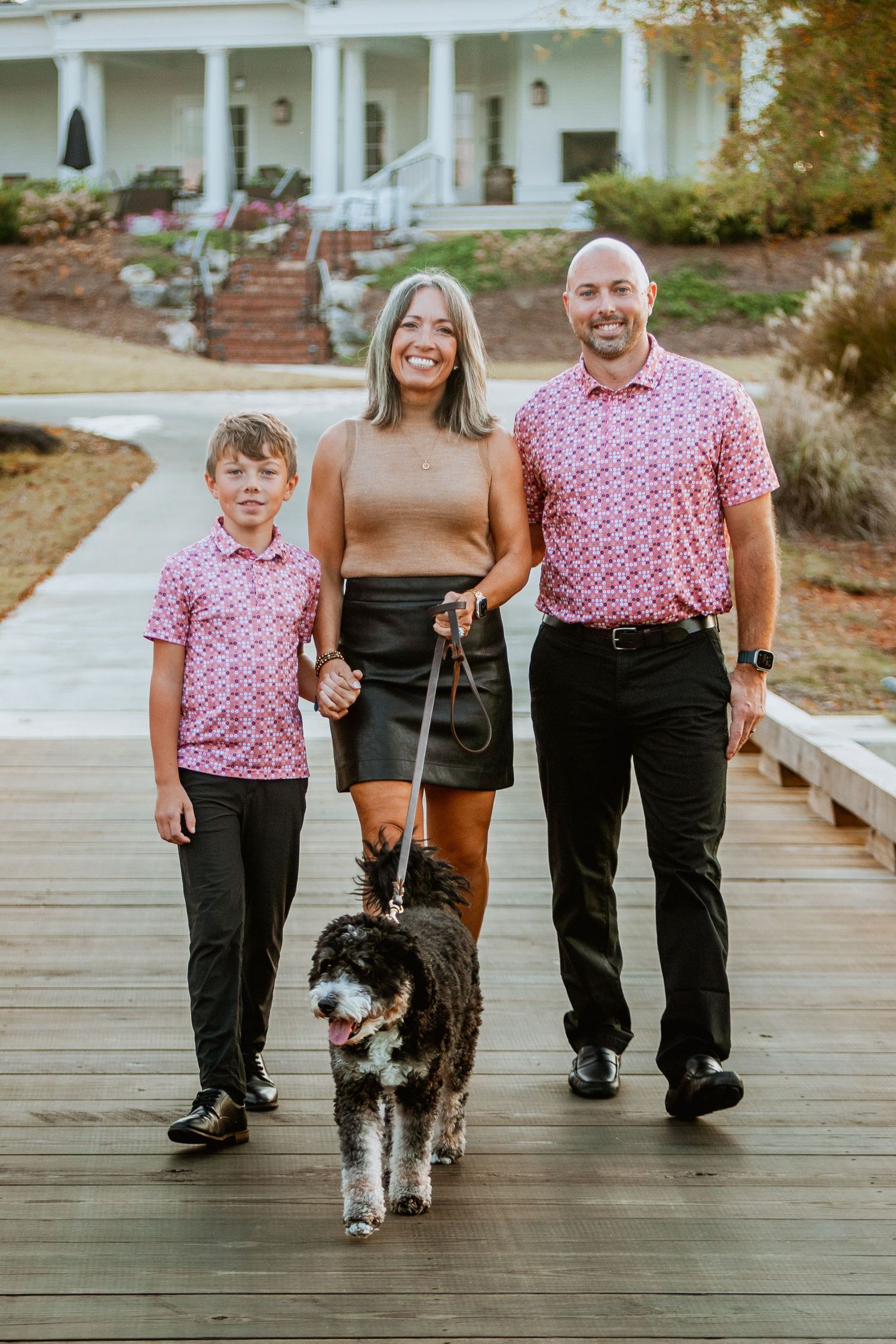 Family of three walks with their bernedoodle dog on wooden bridge during fall photo session.