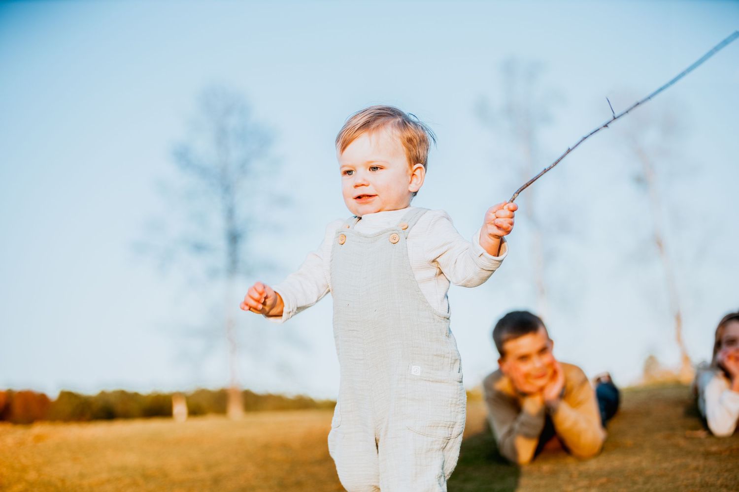 Happy toddler standing on grass waving a stick with siblings relaxing in the background outdoors.