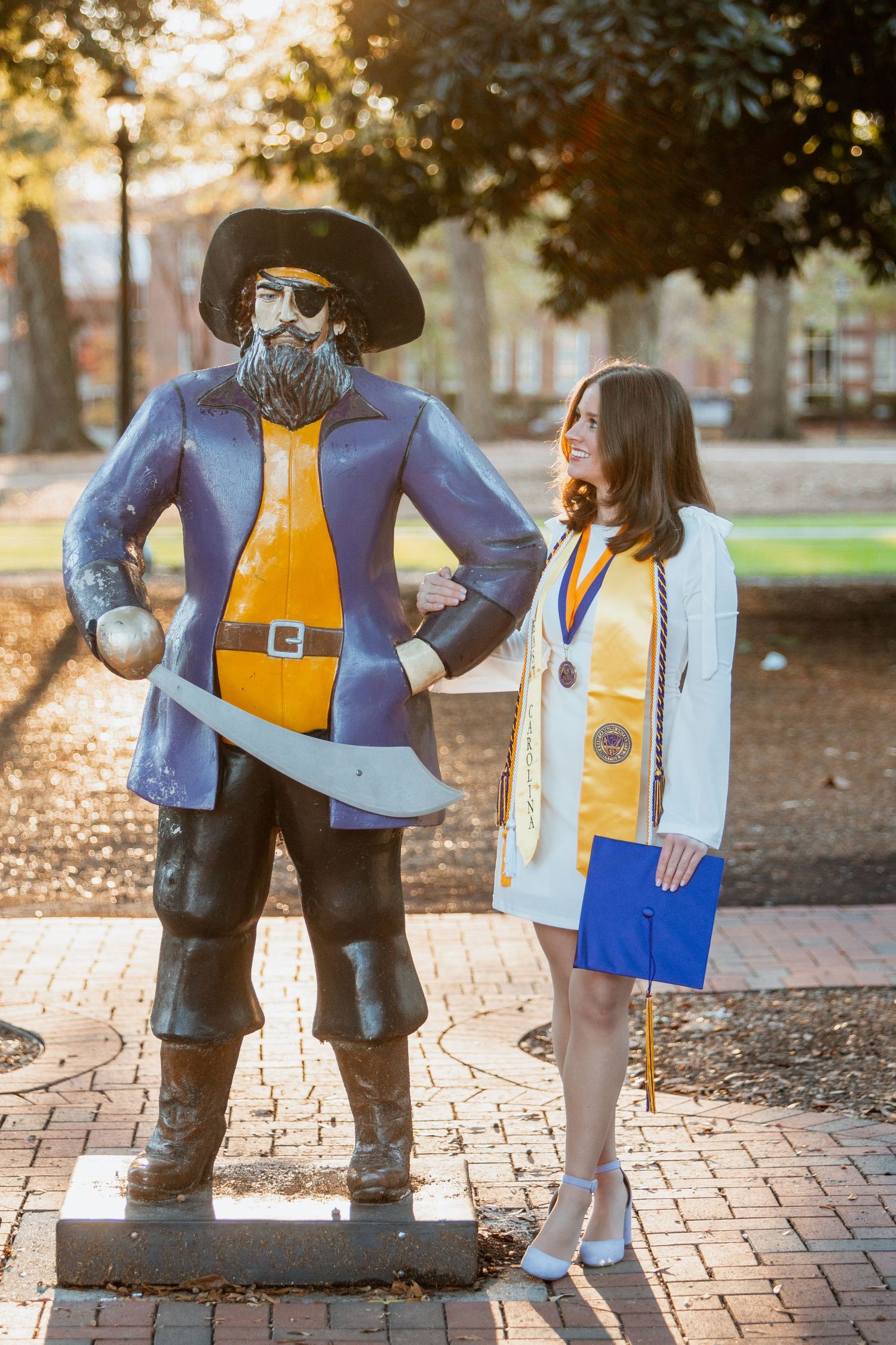 ECU graduate in white dress poses beside Pirate mascot statue holding blue diploma on brick path.