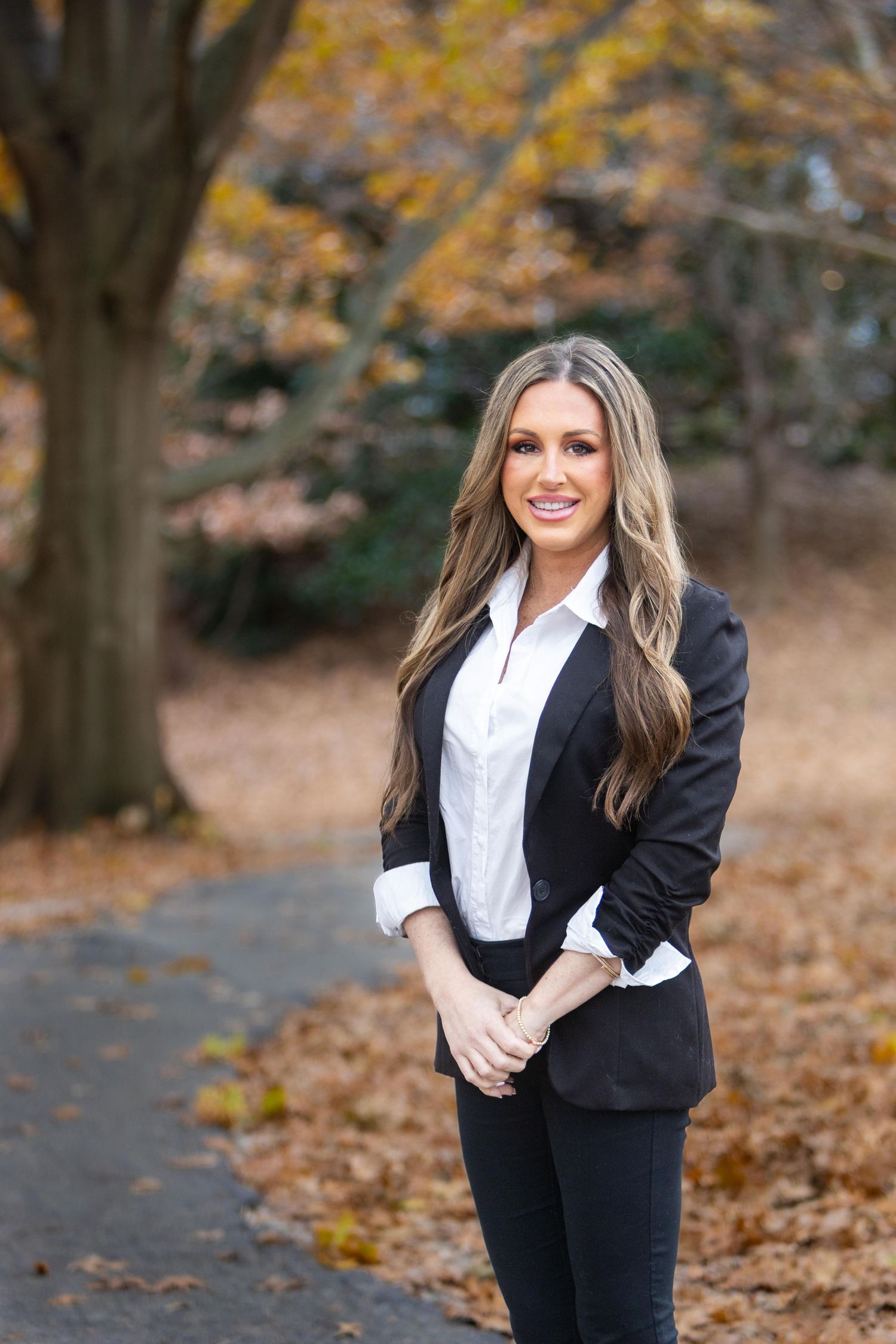 Blonde woman in black blazer and white shirt posing outdoors in autumn park.