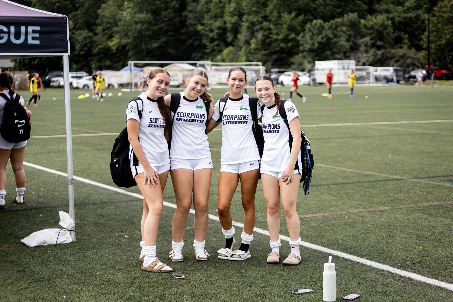 Four female soccer players in white Scorpions uniforms pose together on sideline at National League tournament.