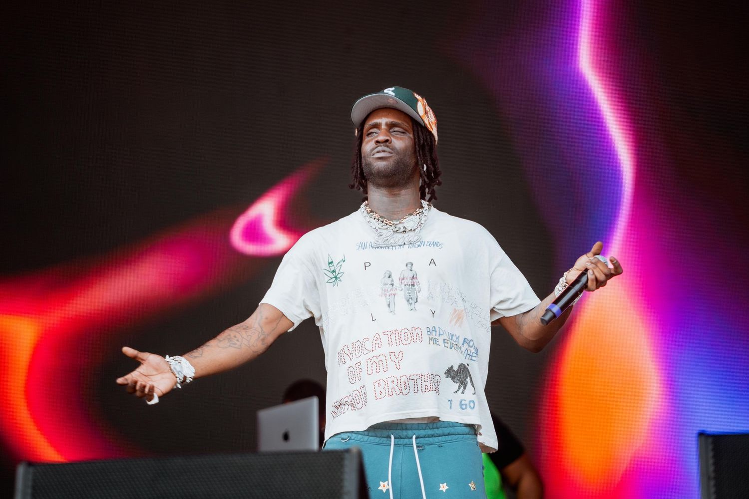 Male rapper on stage in a white graphic tee and teal pants with arms spread wide at an outdoor festival.