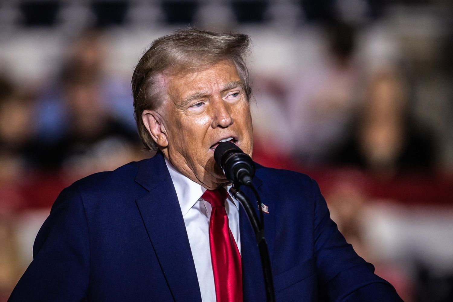 Donald Trump speaks at a microphone during a campaign rally, wearing a navy blue suit and red tie.
