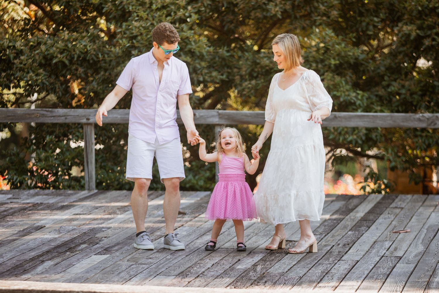 Family of three holding hands on wooden deck with lush green trees in background.