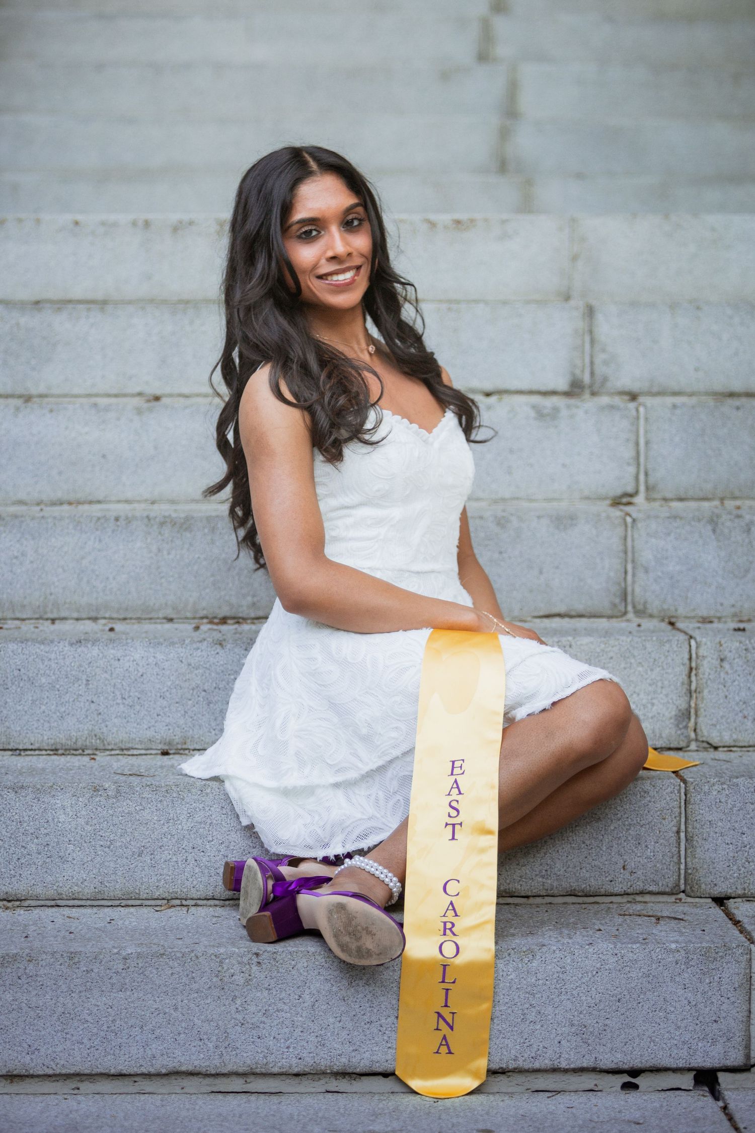 Young woman in white dress sits on stone steps holding an East Carolina University graduation sash.