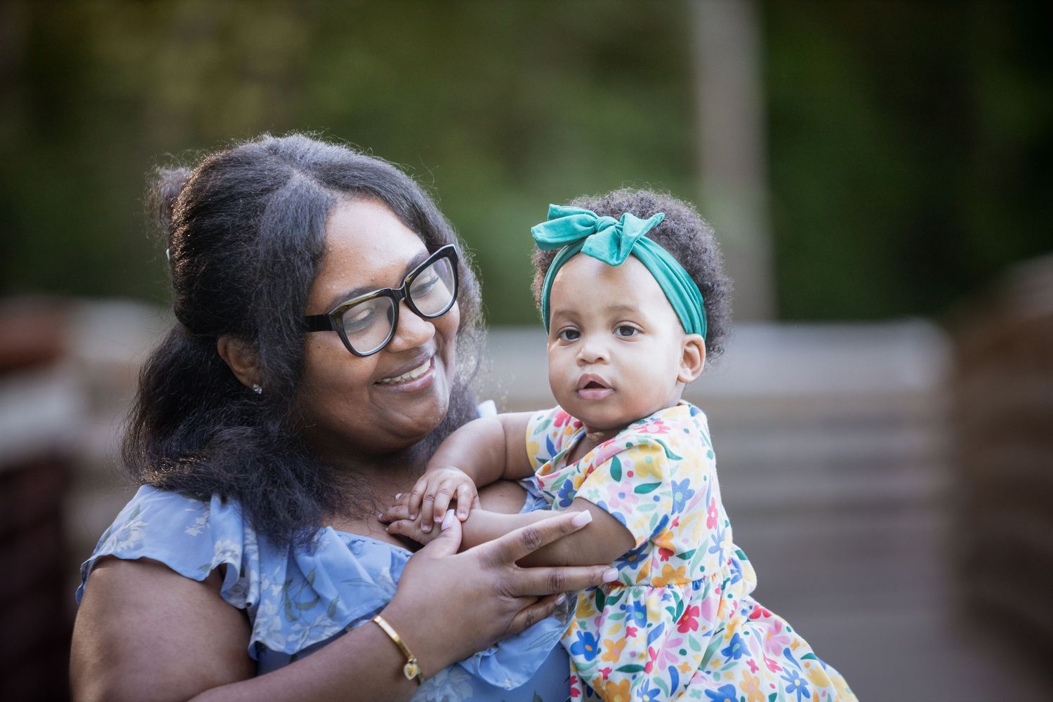 Smiling woman with glasses lovingly holding an alert baby girl wearing a teal bow headband outdoors.
