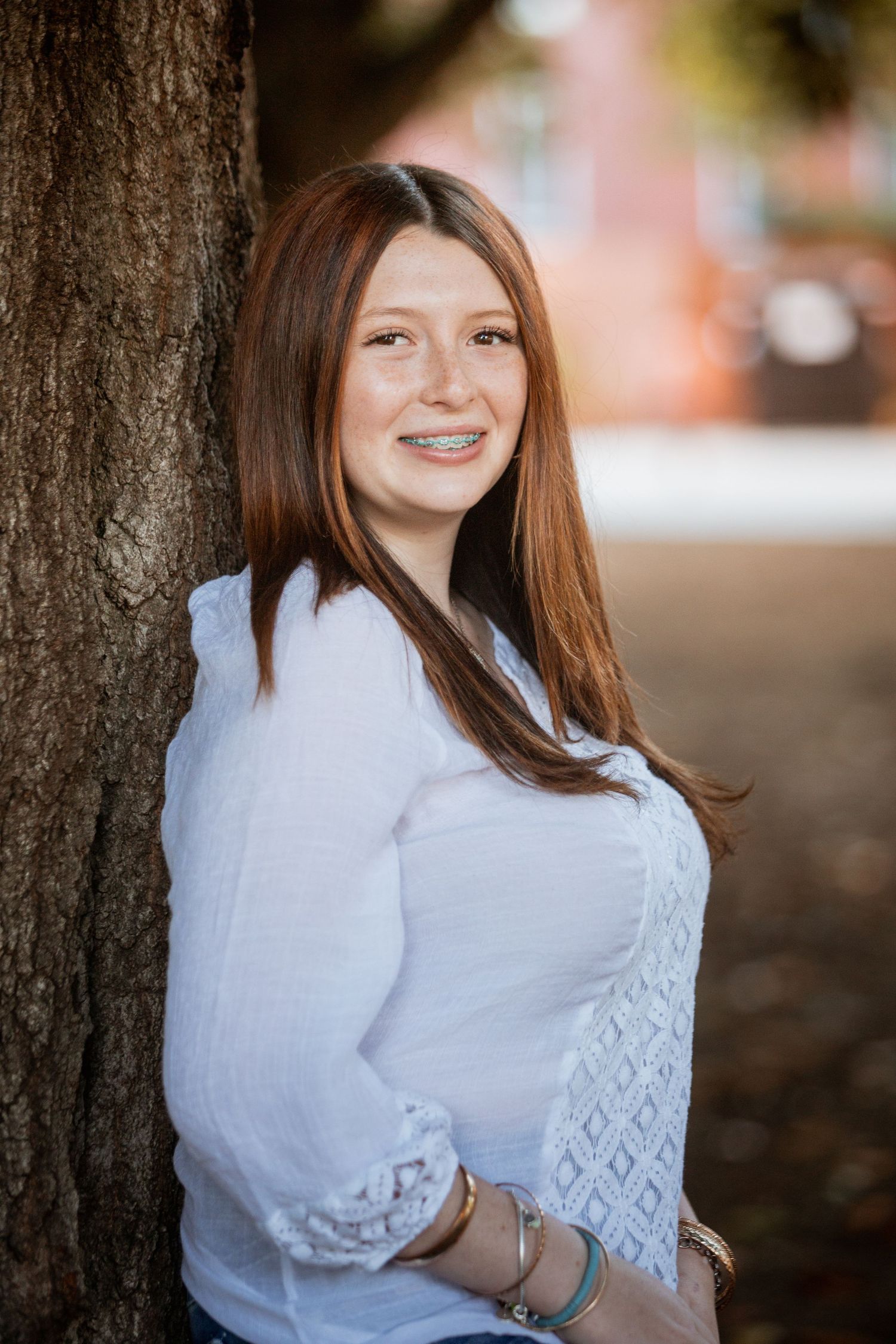 Young woman with red hair and braces smiling against a tree outdoors in a white blouse.