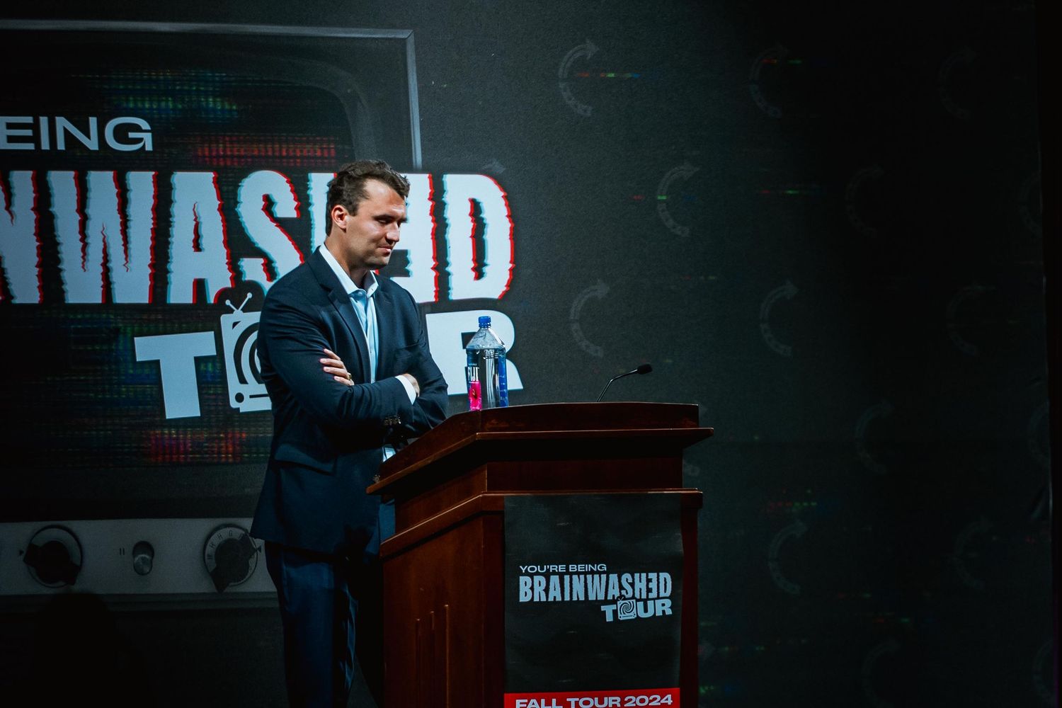 Man in suit standing at the Brainwashed Tour 2024 podium during a speaking event.