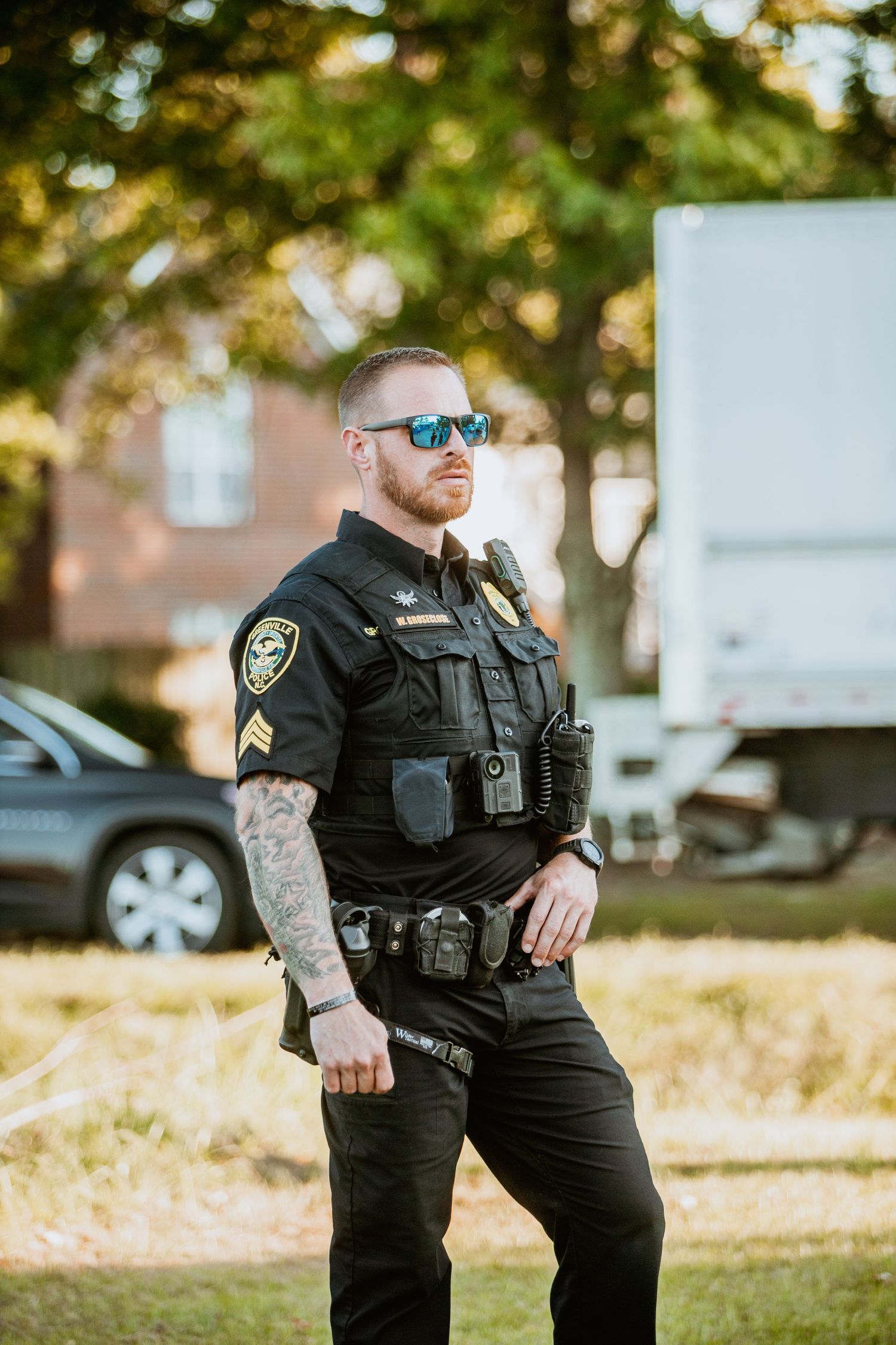 Tattooed police officer in black uniform with sunglasses and badge stands outdoors near parked cars and trees.