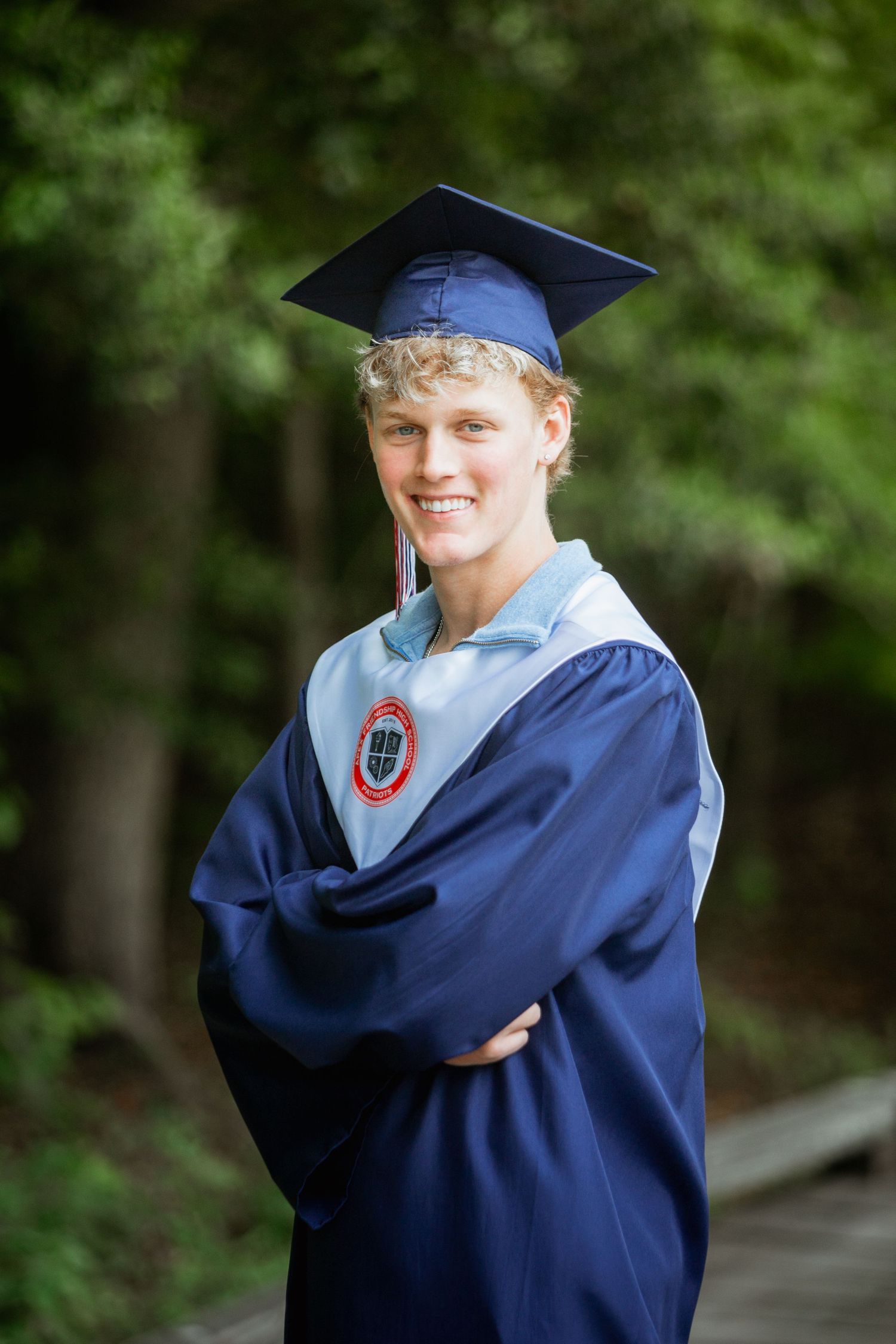 Smiling graduate in navy cap and gown with arms crossed standing in a green forest.