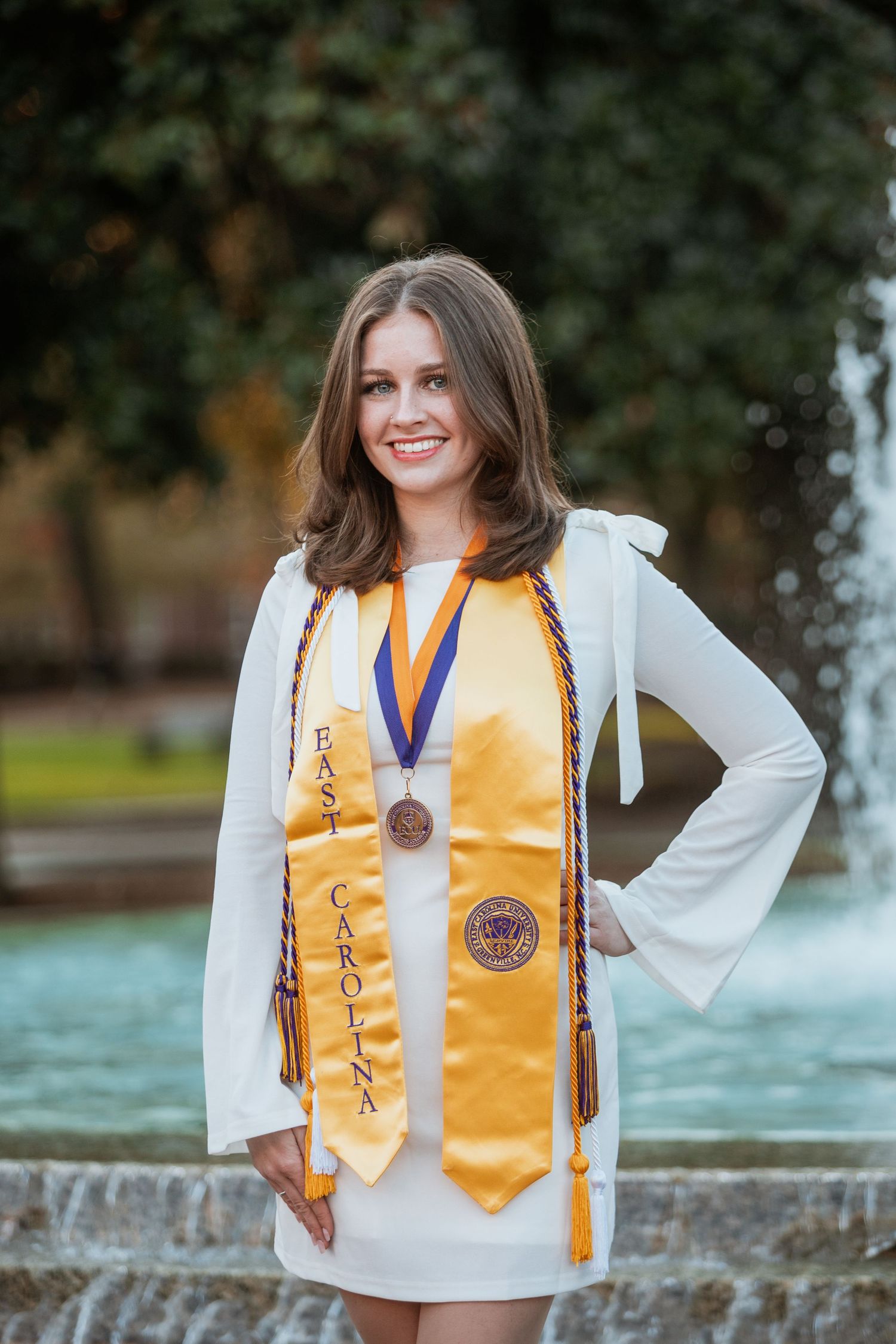 Smiling East Carolina University graduate displays gold honor stole and medal near campus fountain.