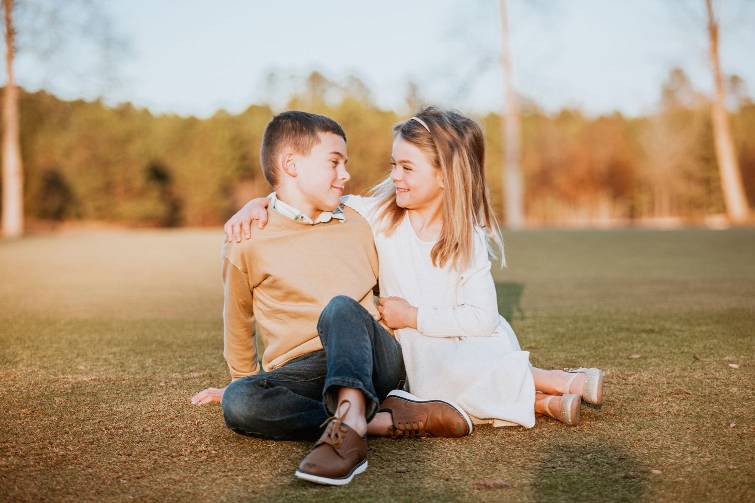 Boy and girl sitting together on green grass, smiling at each other during an outdoor photo session.