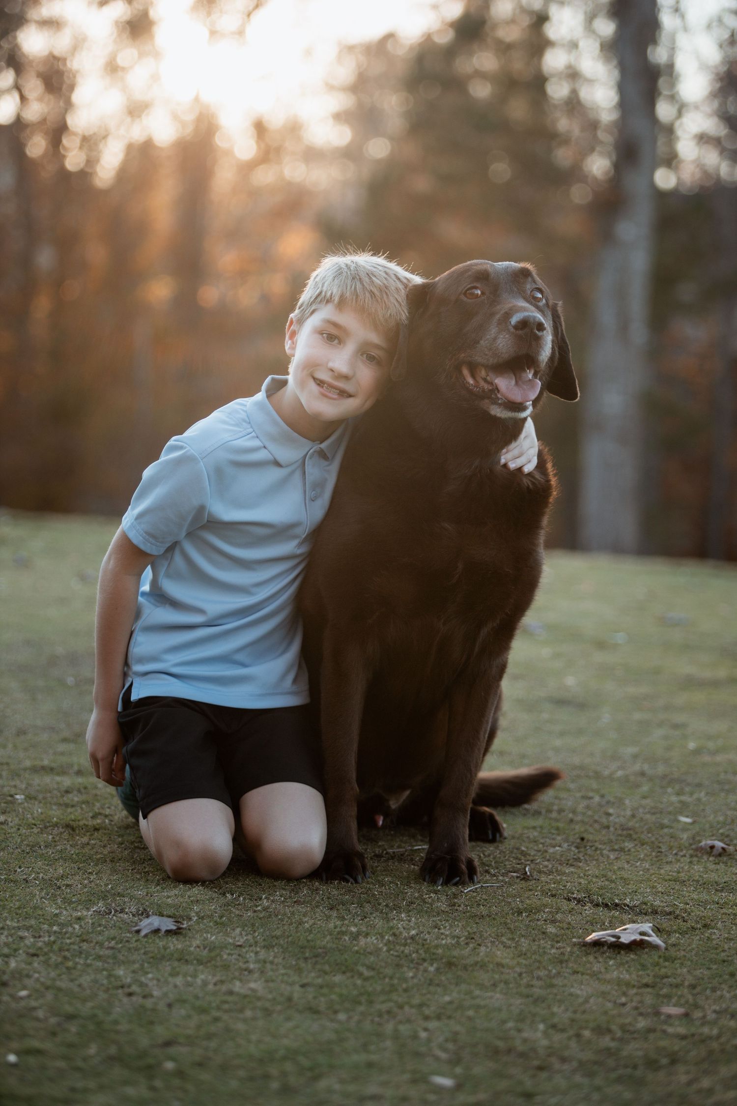 Smiling boy in blue polo shirt hugging a chocolate Labrador dog outdoors at sunset.