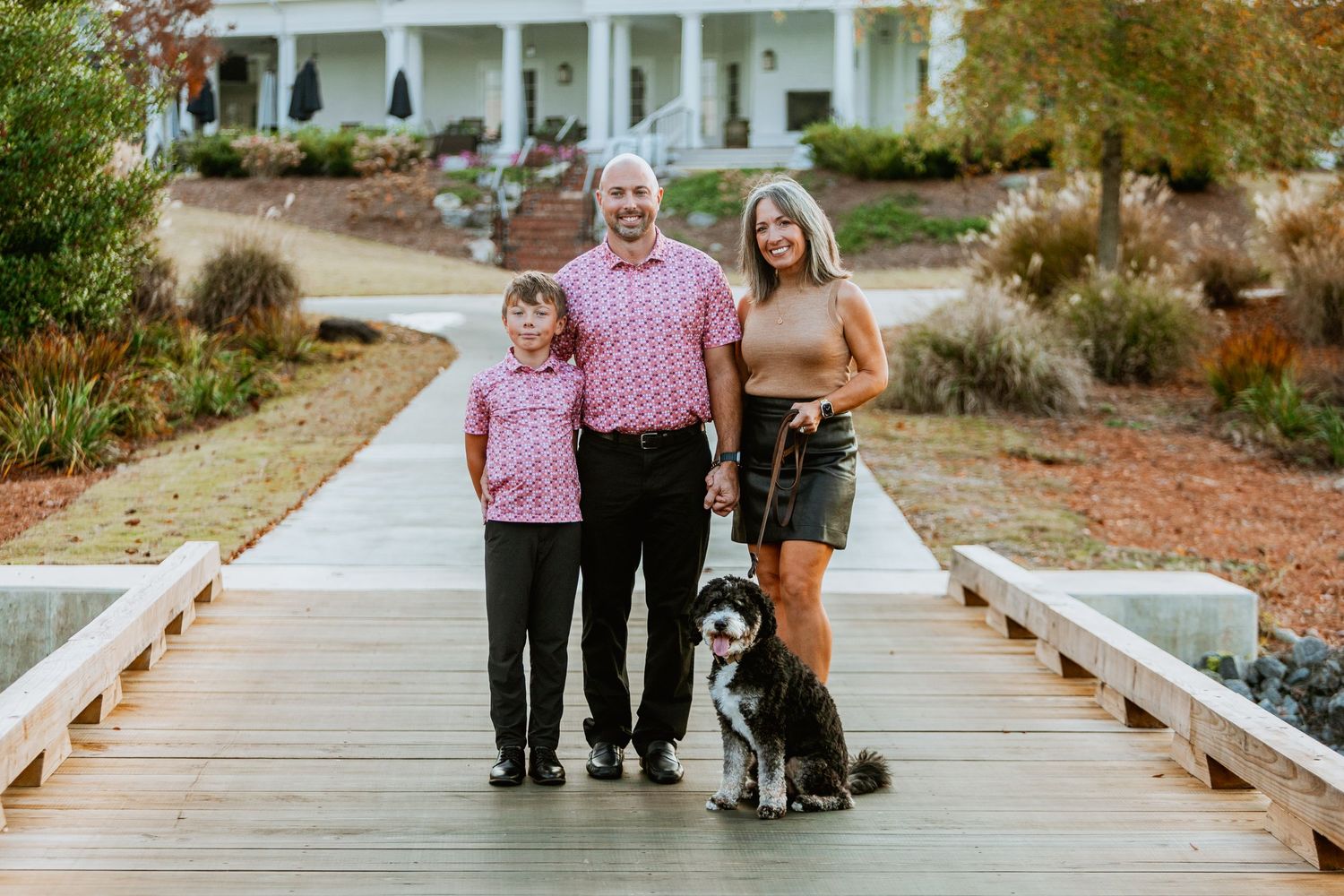 Family of three poses with bernedoodle dog on wooden bridge in front of white estate home.