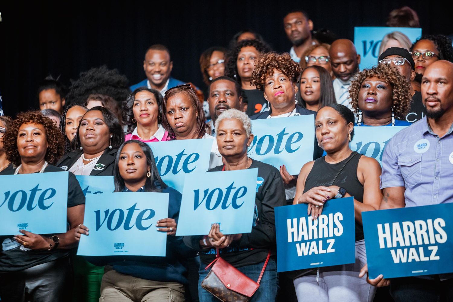 Supporters hold Harris Walz and vote signs at a Kamala Harris campaign rally with a diverse crowd.
