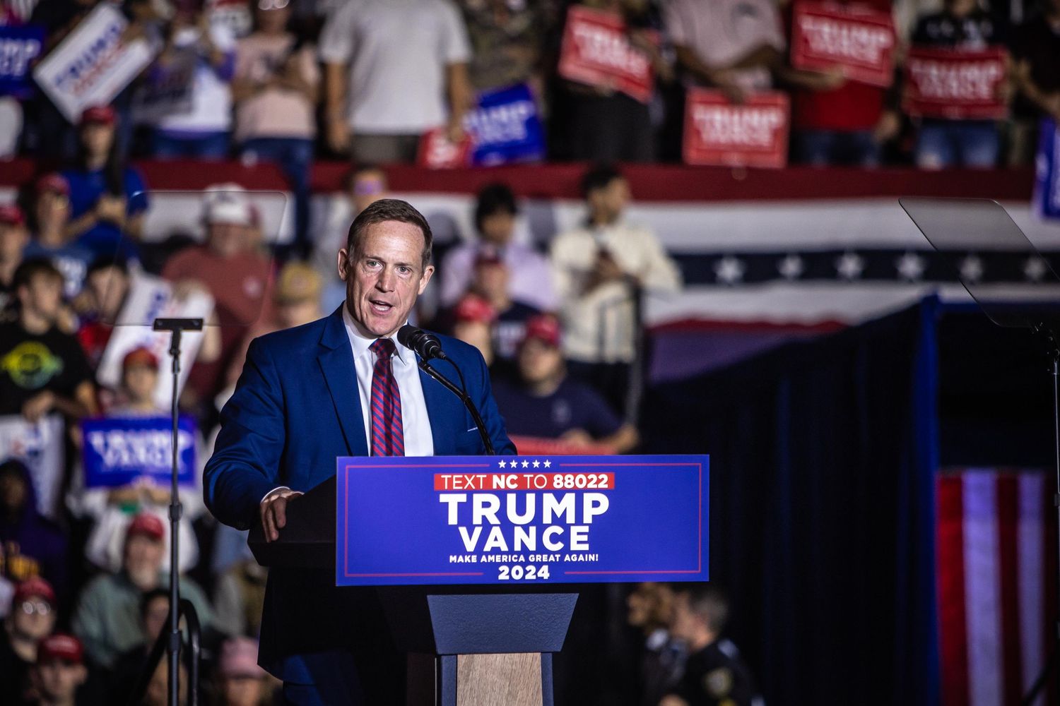 A speaker in a blue suit addresses a large crowd at a Trump Vance 2024 campaign rally in North Carolina.
