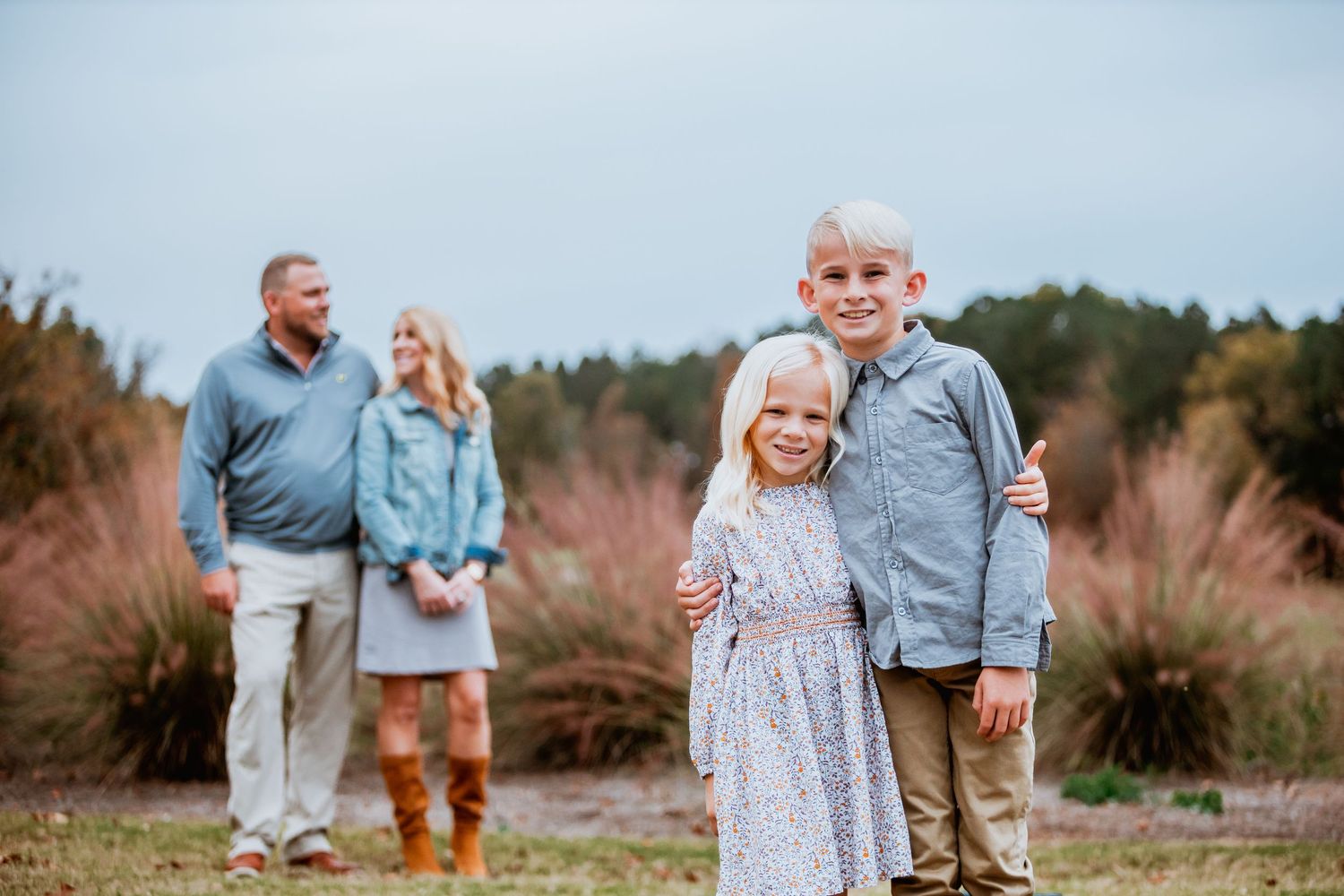 Family of four poses outdoors with children in foreground and parents standing behind among autumn grasses.