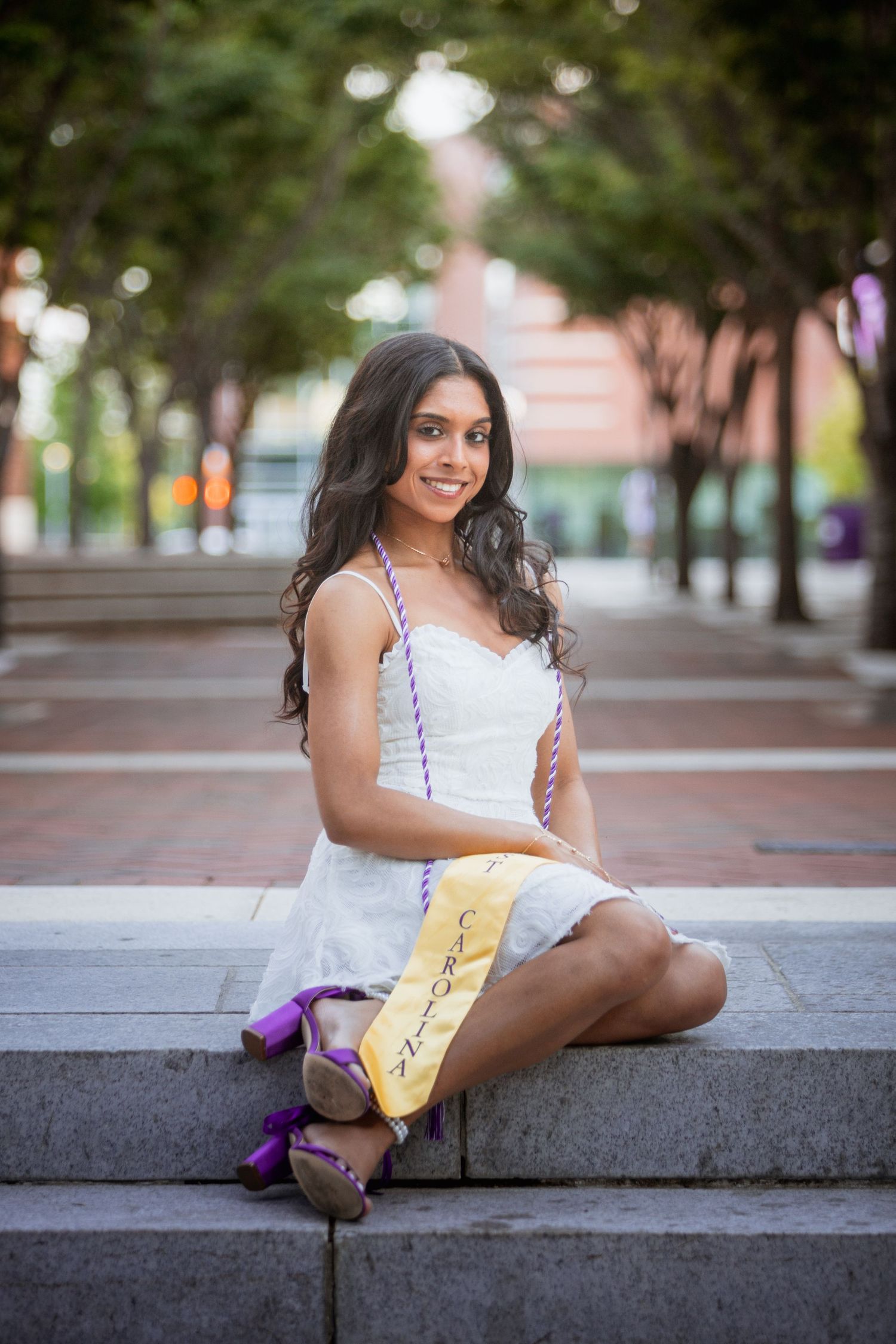 Young woman in white dress with graduation sash sitting on outdoor steps smiling.