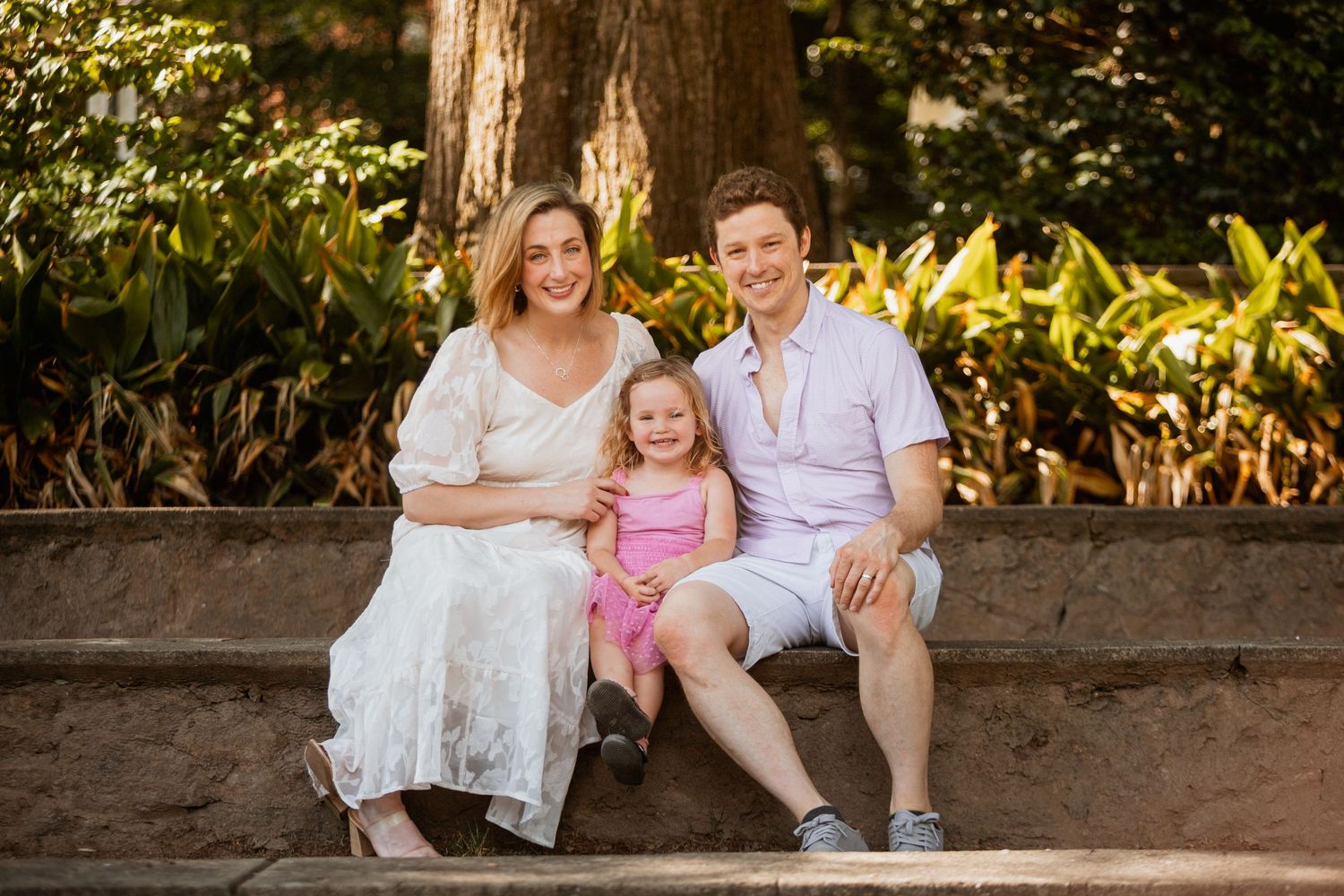 Happy family of three with young girl in pink dress posing on stone steps in park.