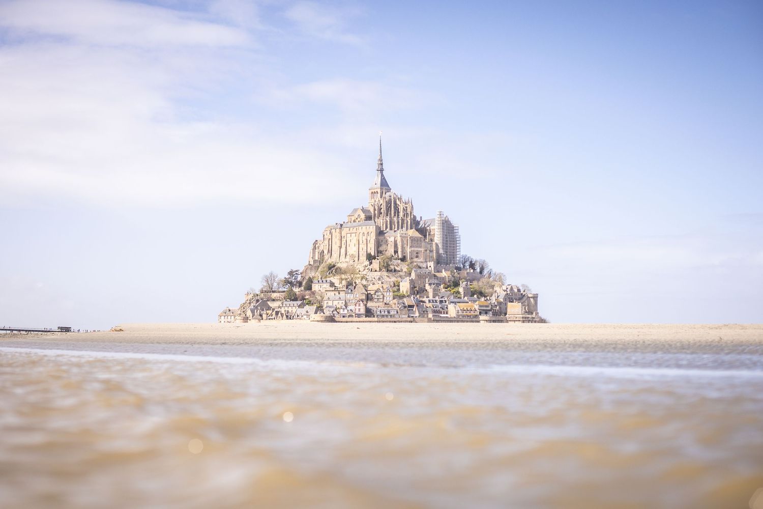 Mont Saint-Michel island abbey reflected in shallow tidal waters under a bright blue sky.