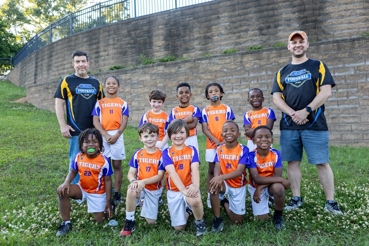 Youth flag football team in orange jerseys poses with two coaches for team photo outdoors.