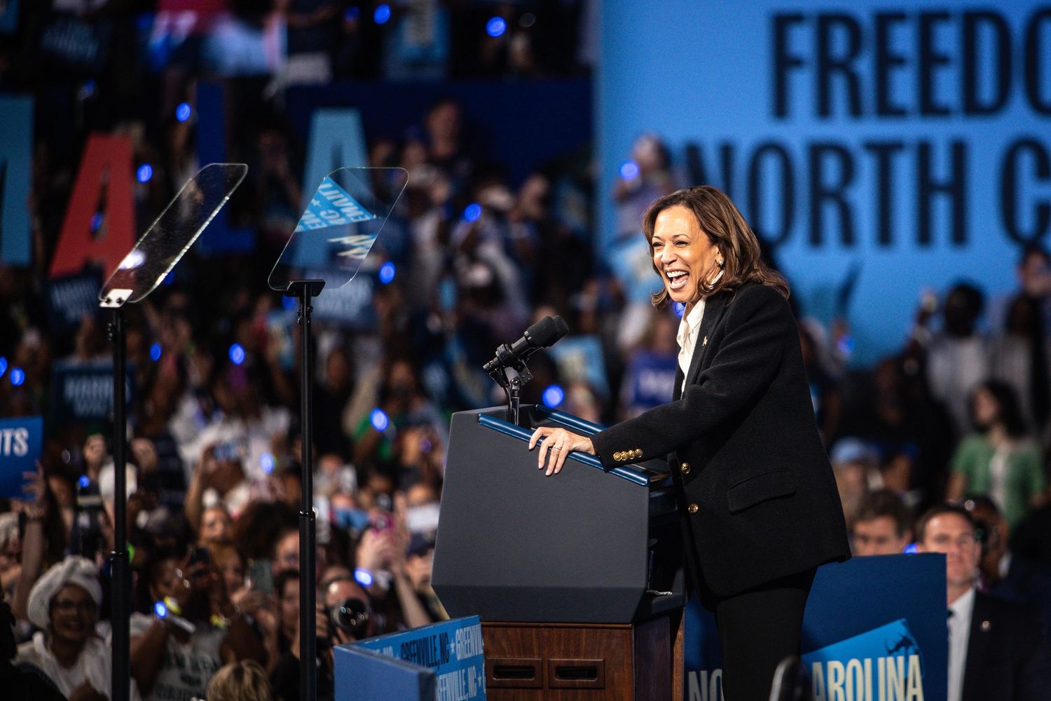 A smiling woman in a black blazer delivers a speech at a Freedom North Carolina campaign rally podium.