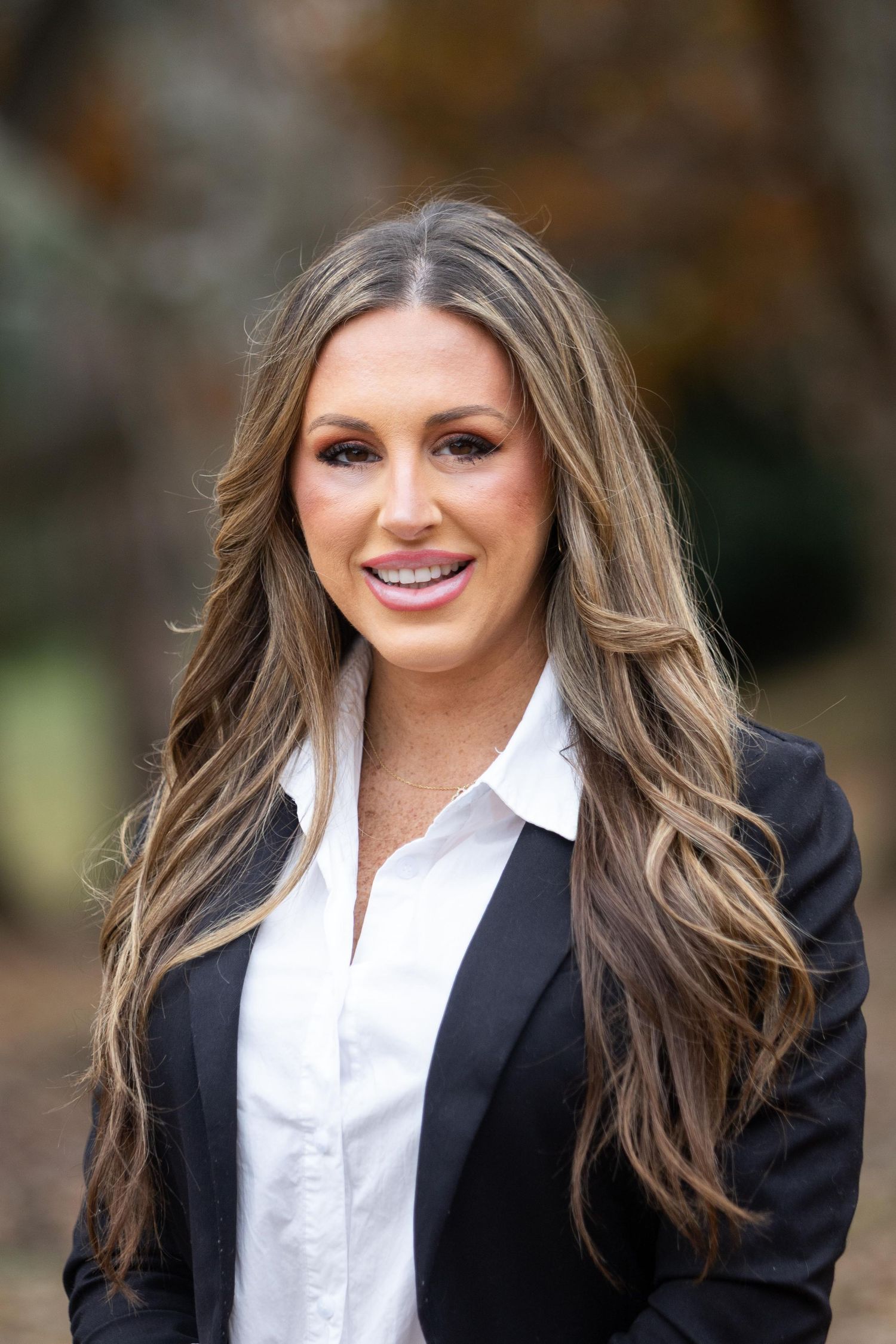 Professional woman with long blonde highlighted hair wearing a black blazer and white shirt smiling outdoors.
