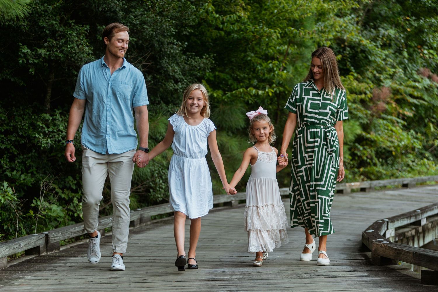 Family of four holding hands walking on a wooden boardwalk surrounded by lush green trees.