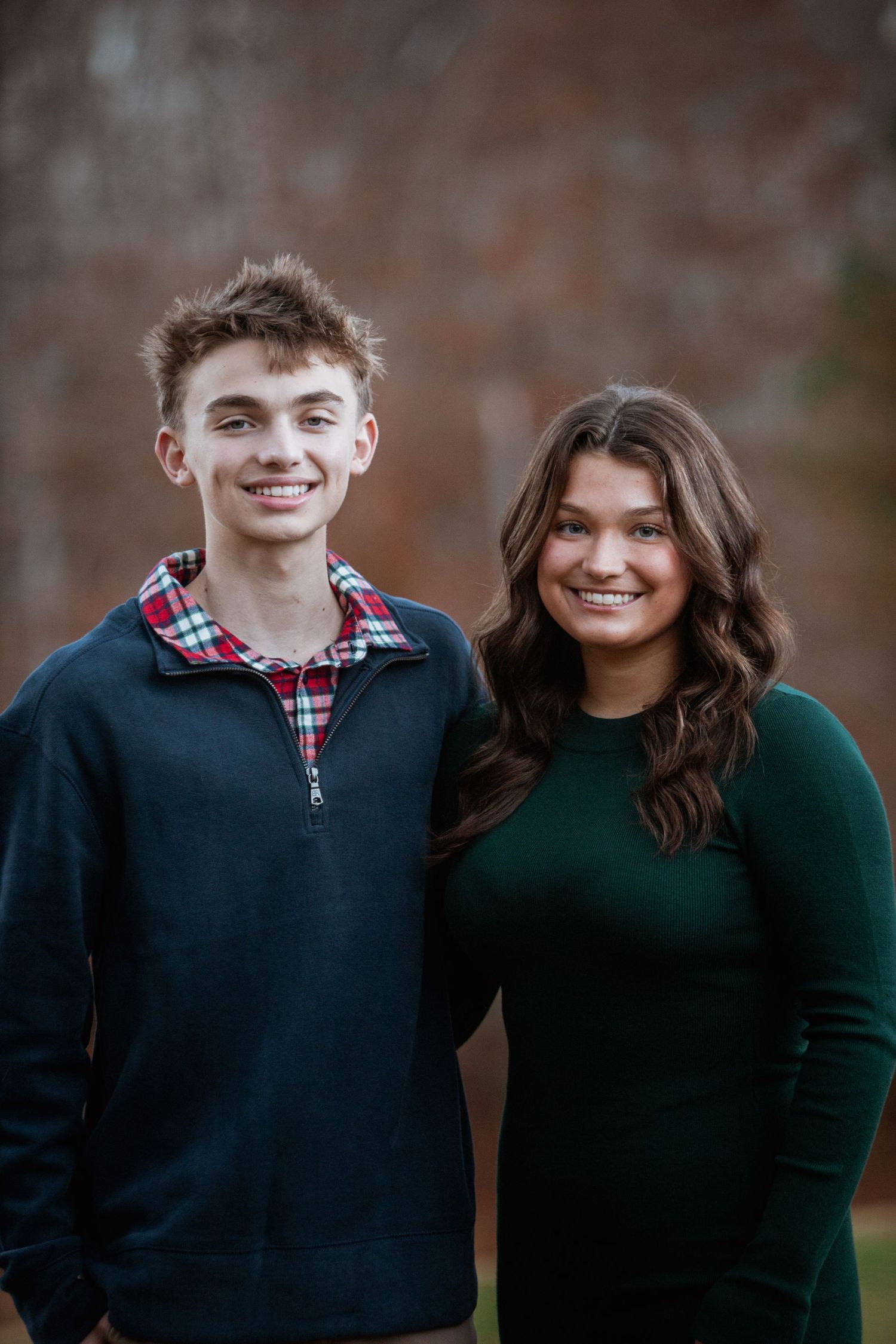 Teen boy in navy pullover and girl in green dress smile together in front of a brick wall.