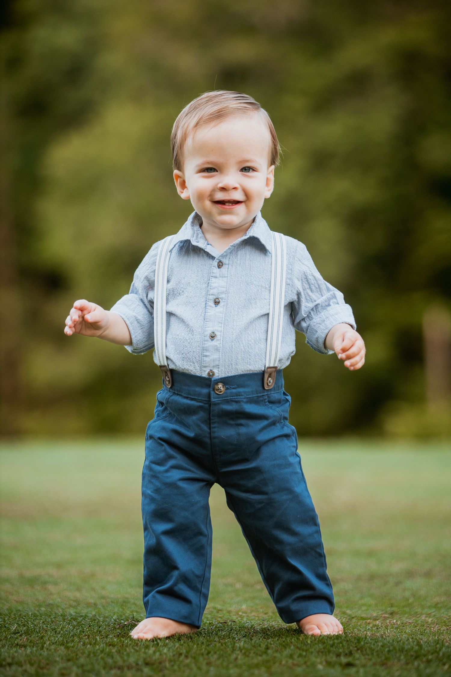 Adorable smiling toddler boy in blue shirt and suspenders standing barefoot on green grass outdoors.