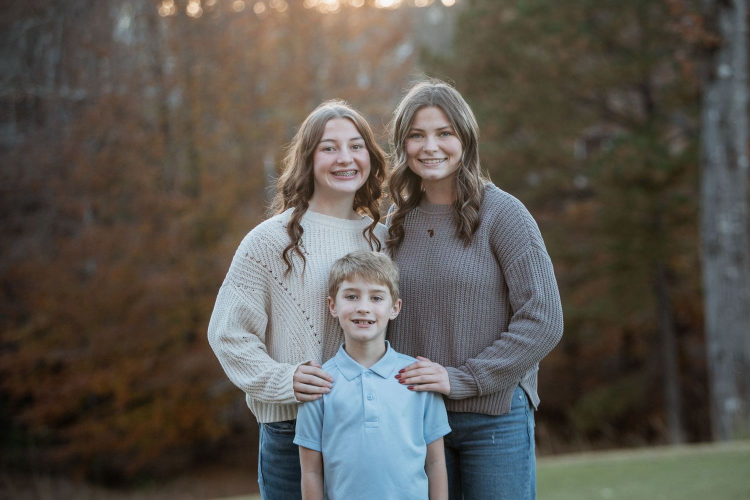 Two teenage girls and a young boy posing together for a sibling portrait in an autumn outdoor setting.