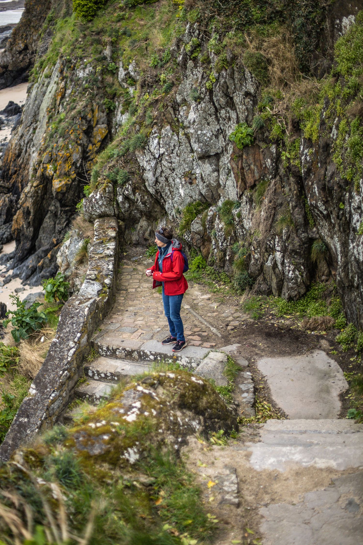 Woman in red jacket standing on stone steps carved into rocky coastal cliffs on a hiking trail.