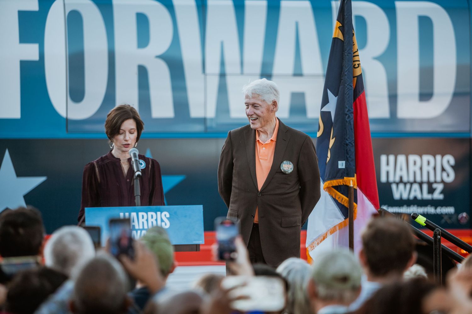 Two people stand on stage at a Harris Walz Forward campaign rally with North Carolina flag.