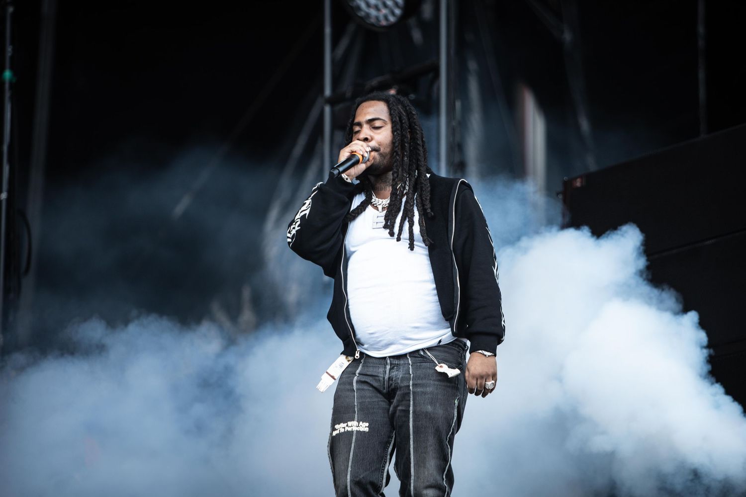 Hip-hop artist performing on stage in a black jacket amid dramatic smoke effects at an outdoor concert.